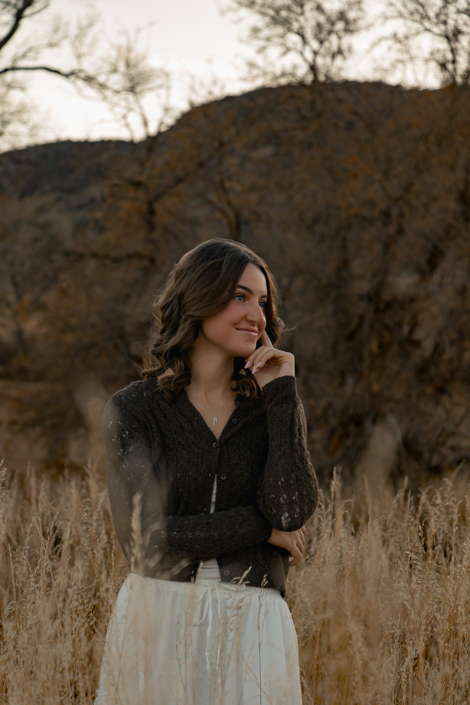 Person in black top and white skirt standing in autumn field at sunset with mountains behind.