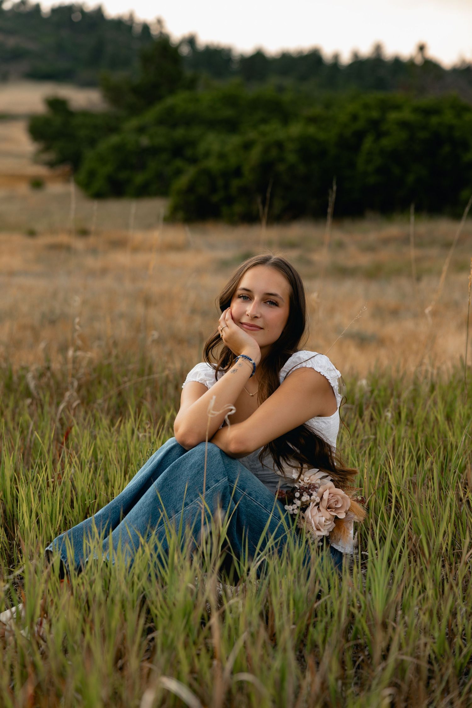Young woman sitting in tall grass field, wearing white top and jeans, smiling softly with hand on chin at sunset.