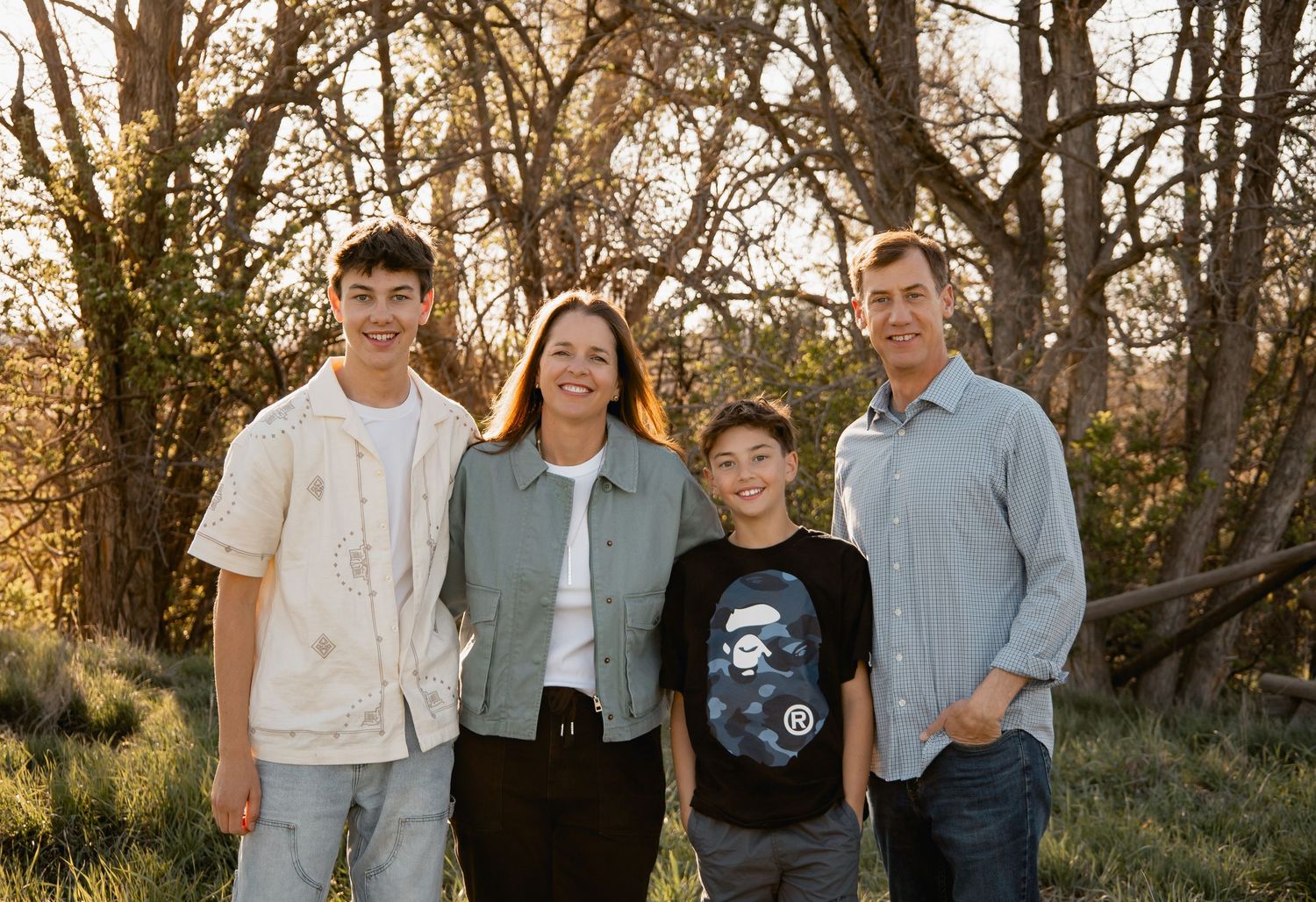 Family stands together smiling in a natural outdoor setting with wooded backdrop during golden hour.