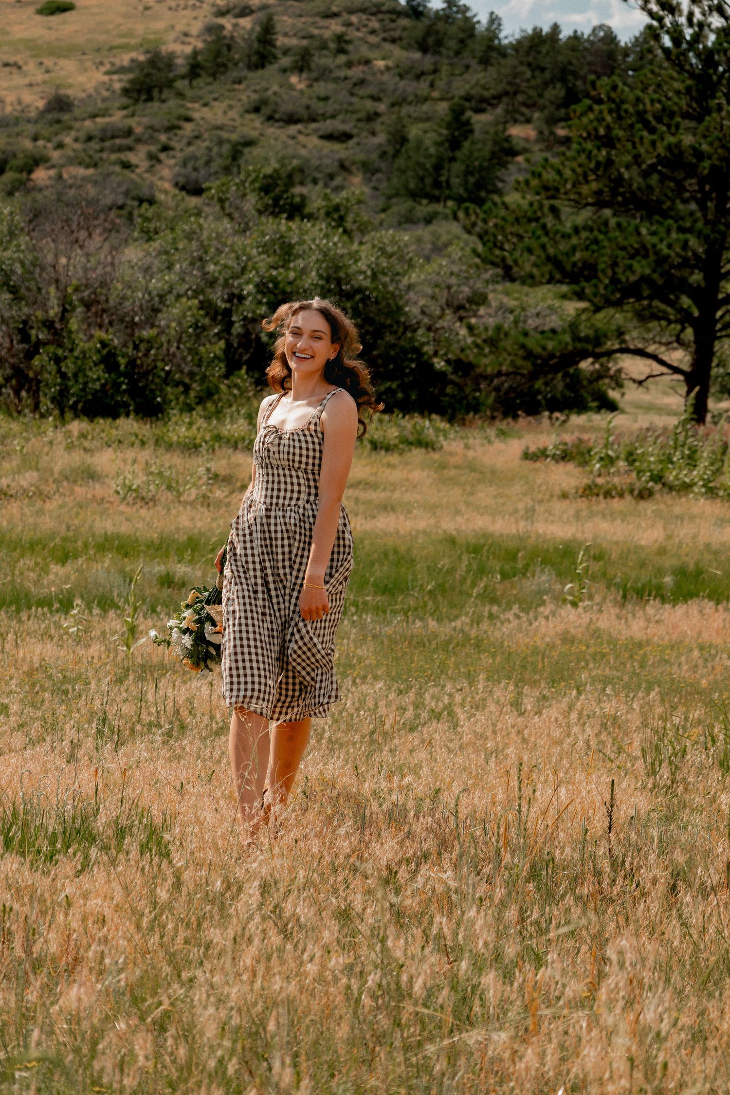 Person in gingham dress walks through tall grass in a sunny meadow surrounded by rolling hills and trees.