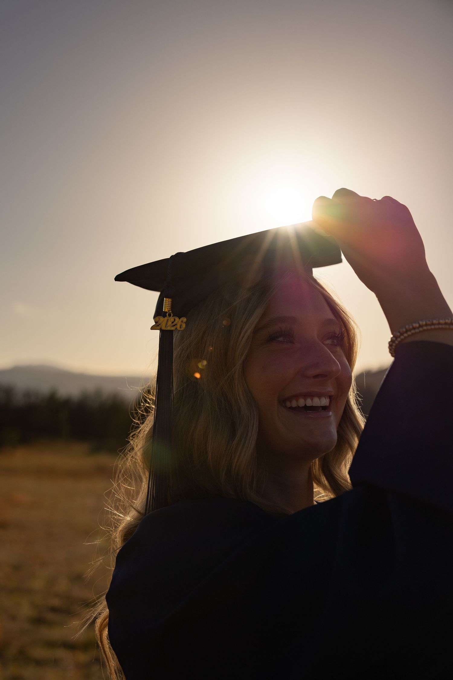 Smiling graduate holding black mortarboard cap outdoors at golden sunset with lens flare and mountain backdrop.