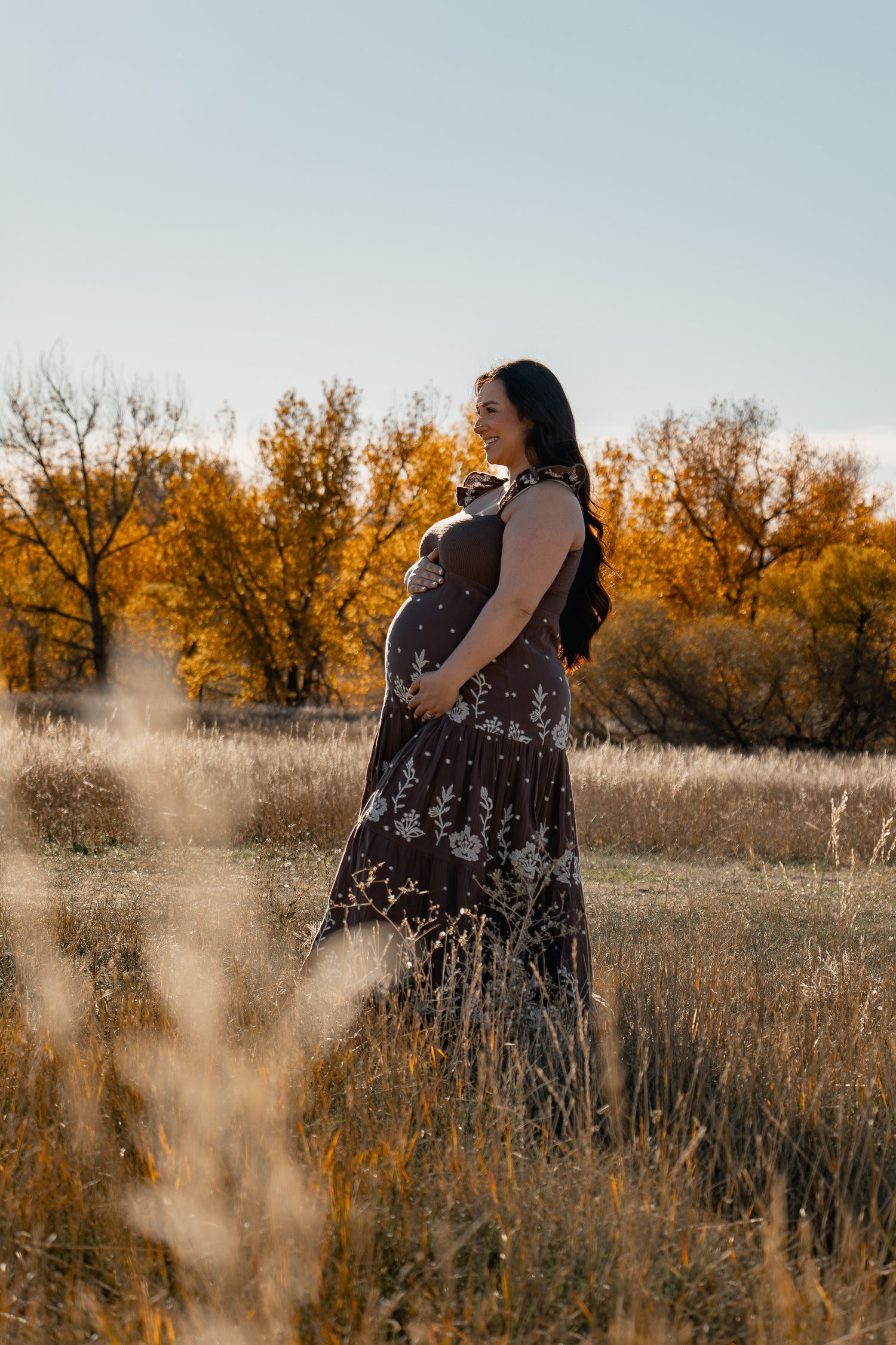 A figure in a dark floral dress stands in a golden autumn field at sunset with tall grasses in the foreground.