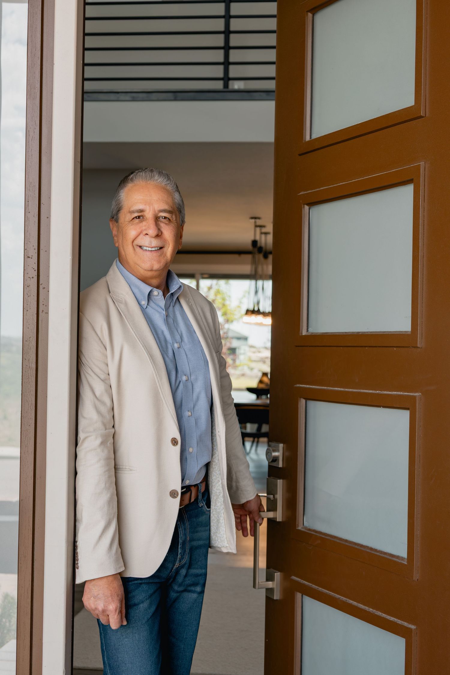 A person in a beige blazer and blue shirt smiles warmly while standing in a modern doorway at home.