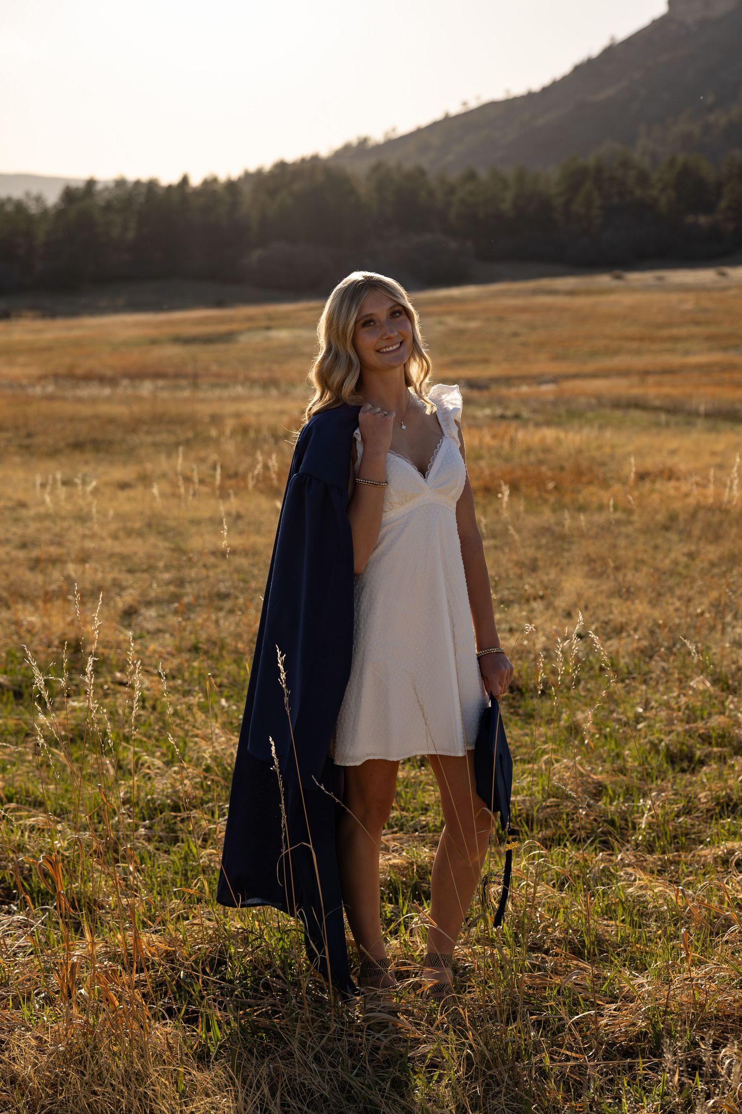 Smiling blonde woman in white dress holding navy blazer standing in golden field at sunset with mountains in background.
