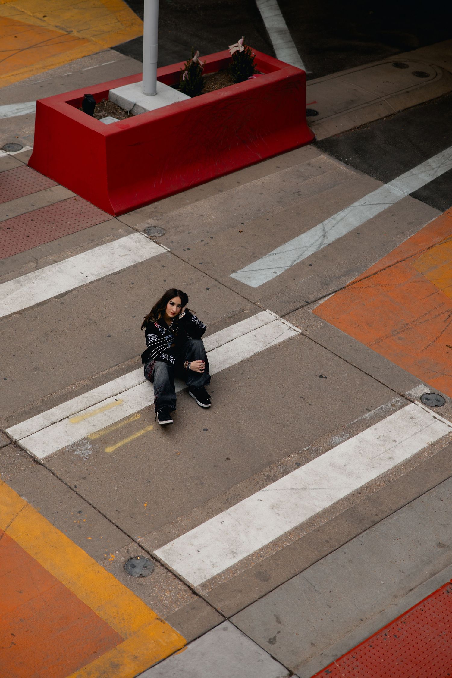 Aerial view of a person sitting on a crosswalk surrounded by colorful urban pavement markings and a red planter.