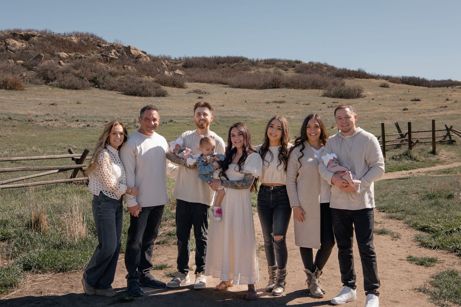 A group in casual white outfits smiling together in front of a desert landscape with wooden fencing and hills.