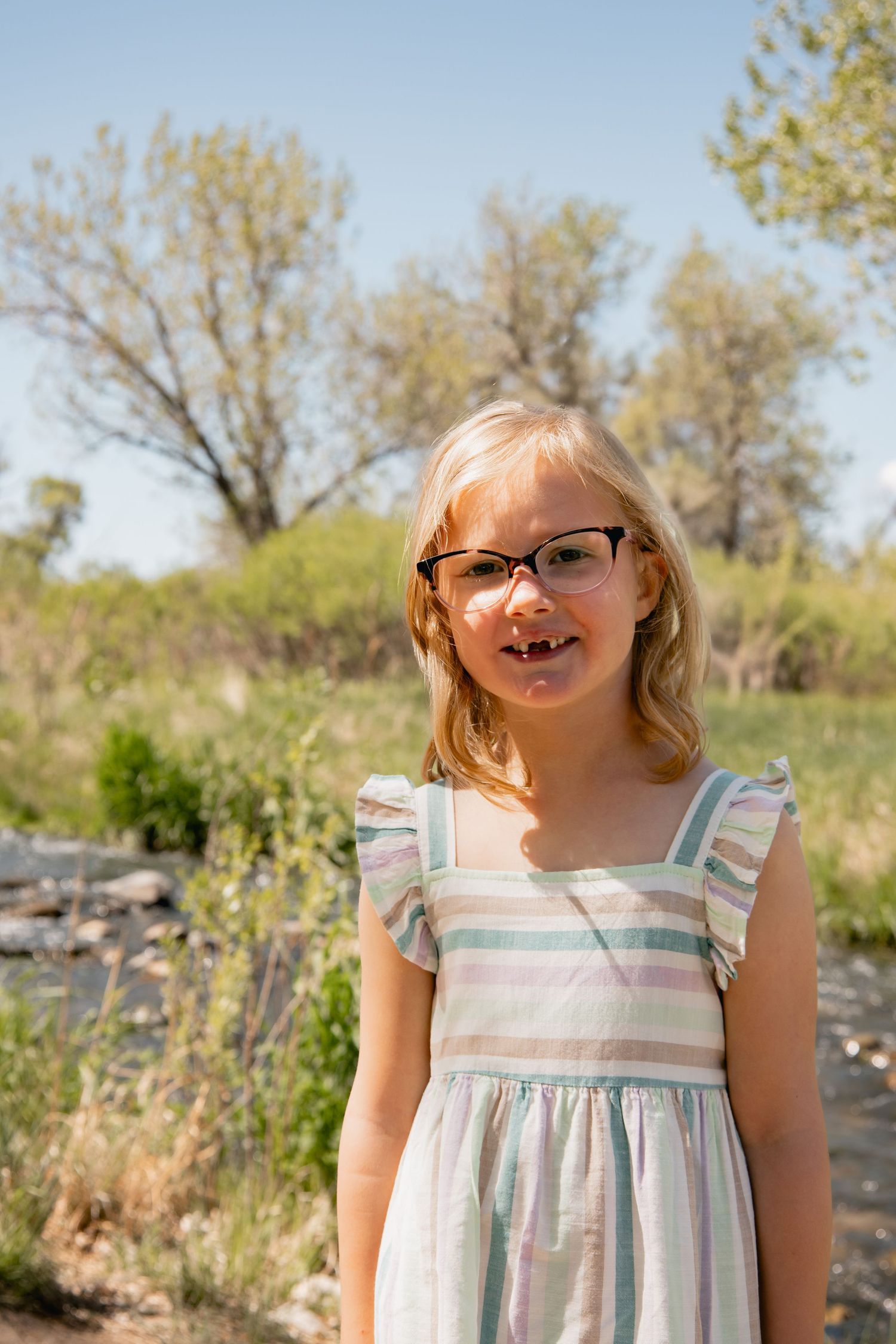 A child wearing glasses and a striped dress smiles while standing outdoors on a sunny day.
