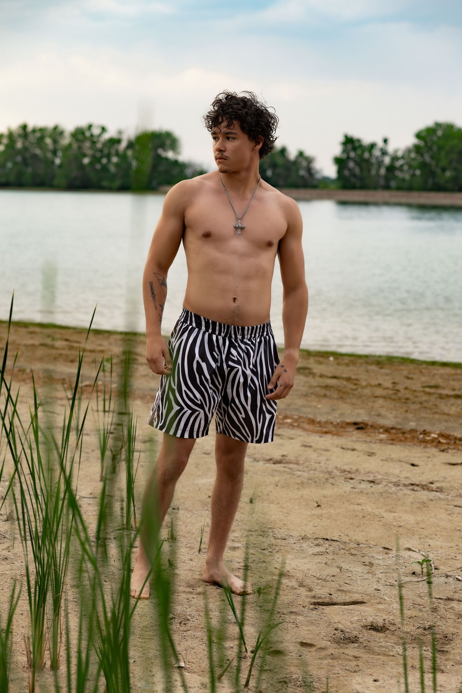 Shirtless young man in zebra print swim shorts standing barefoot on sandy beach by calm lake.
