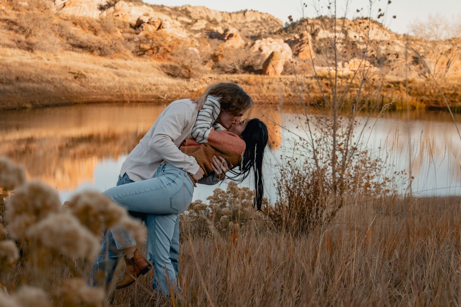 A couple shares a romantic moment at sunset by a tranquil lake surrounded by golden autumn grass and reeds.