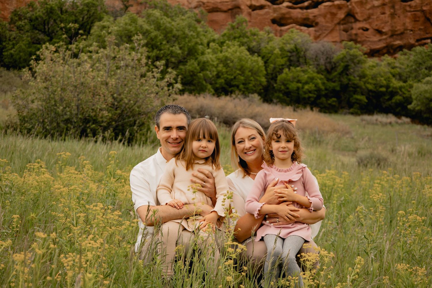 A family sits together in a wildflower meadow with red rock formations and green trees in the background.