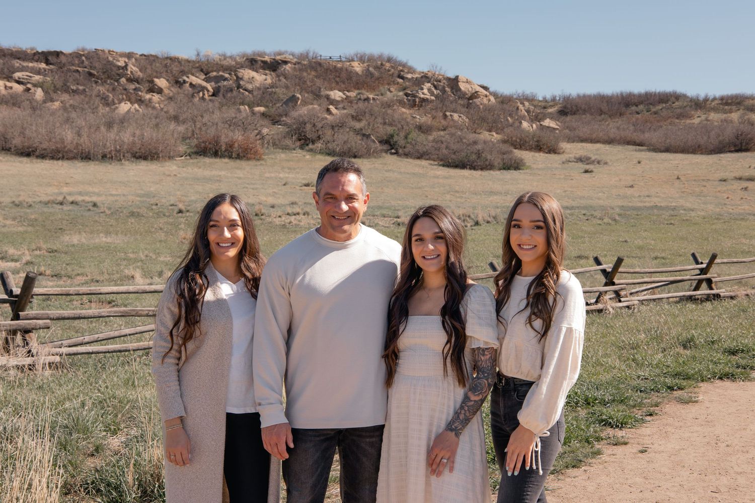 A group of people in coordinating white and neutral outfits pose together on a dirt path with a rustic wooden fence behind them.