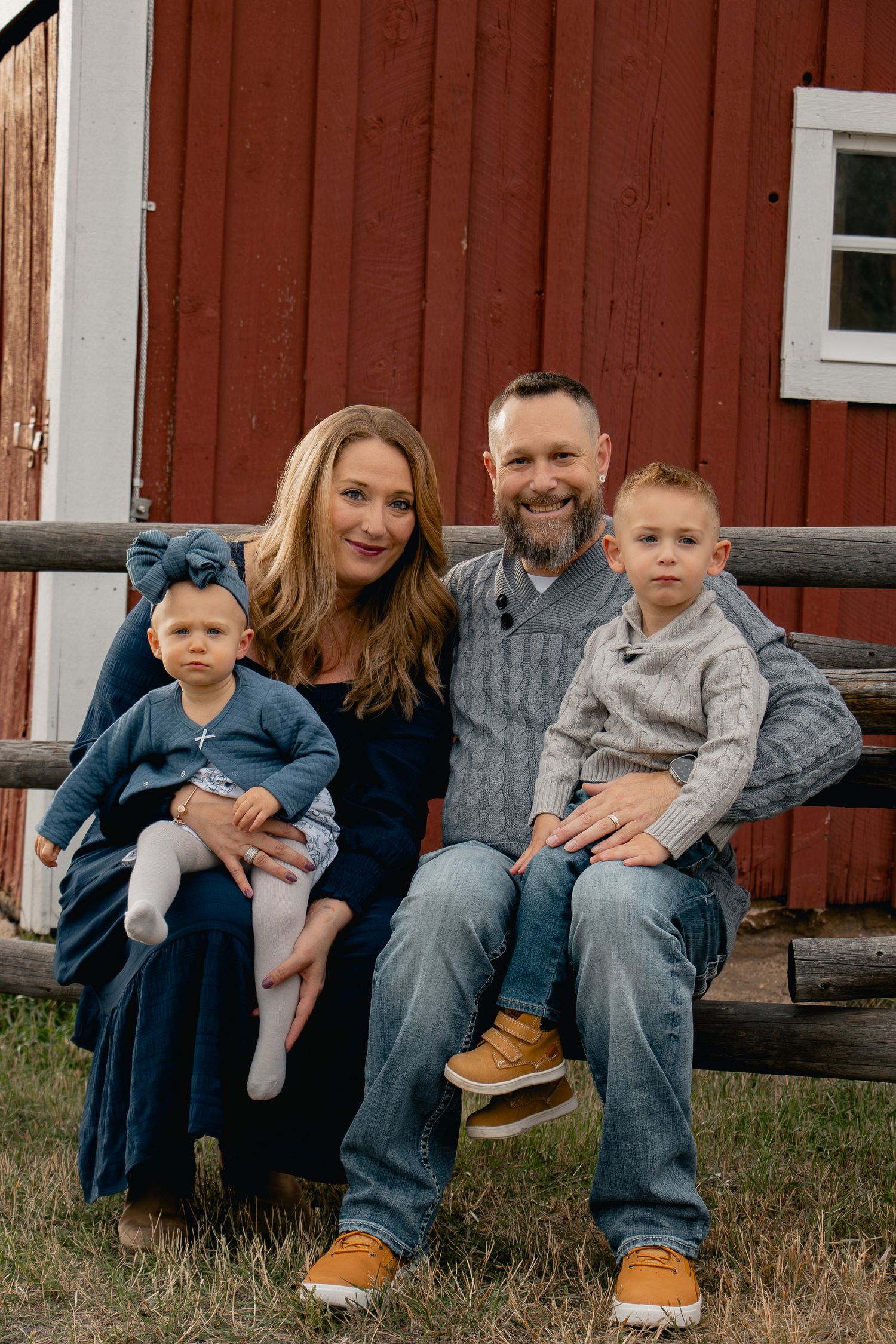 A family sits together on wooden steps in front of a rustic red barn on a sunny day.