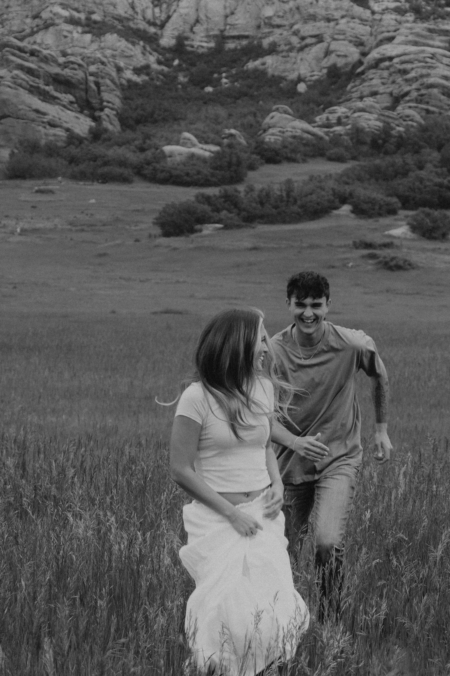 A couple shares a candid moment running through a tall grass meadow with rocky cliffs in the background.