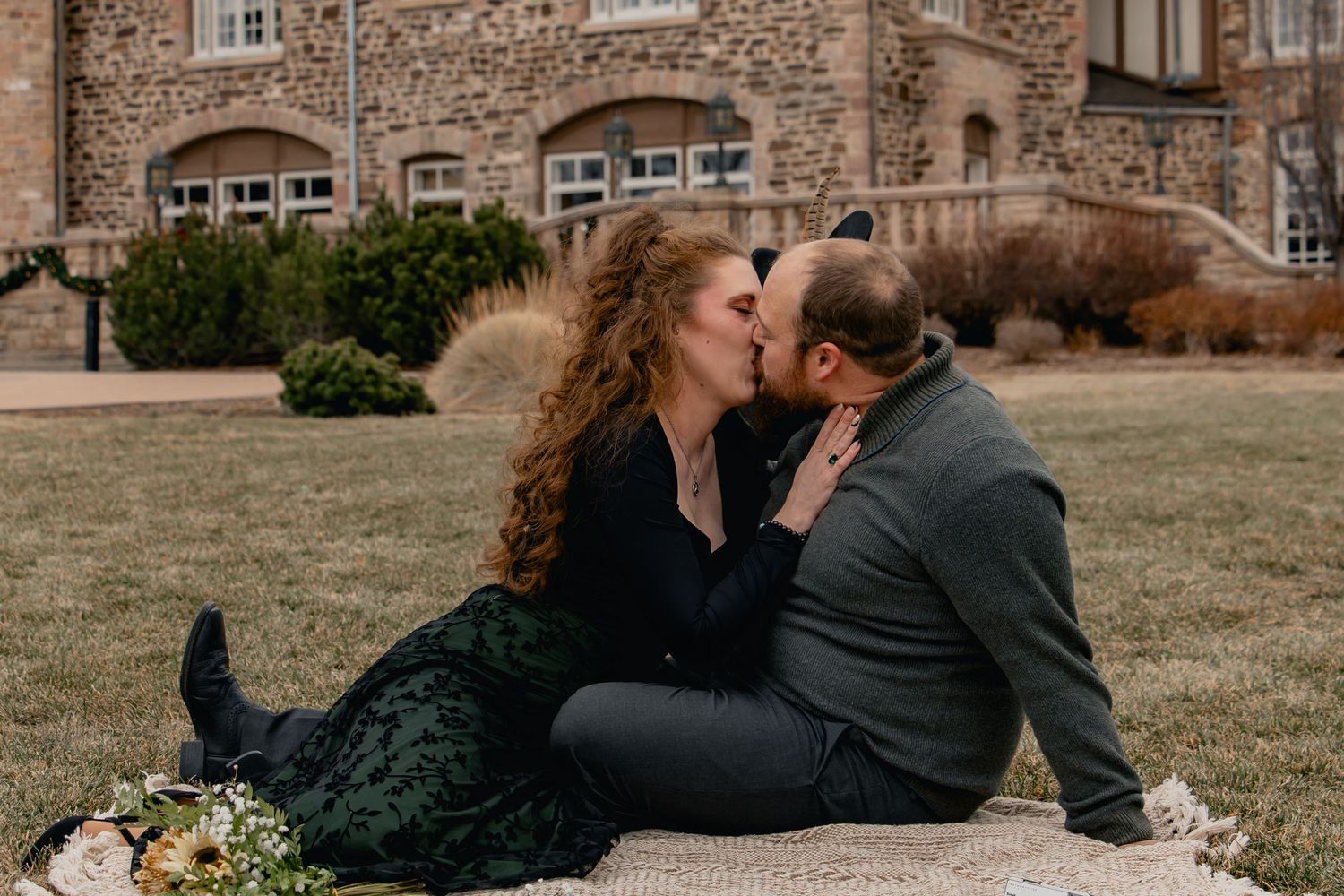 A couple shares a romantic kiss while sitting on a blanket with flowers outside a stone castle building.