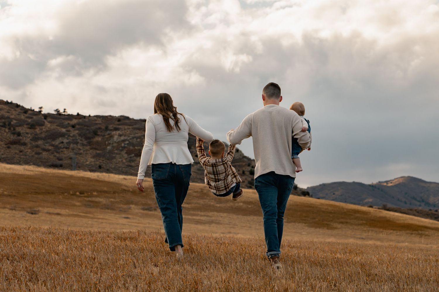 A couple walking hand in hand through a golden grass field with mountains in the background at sunset.