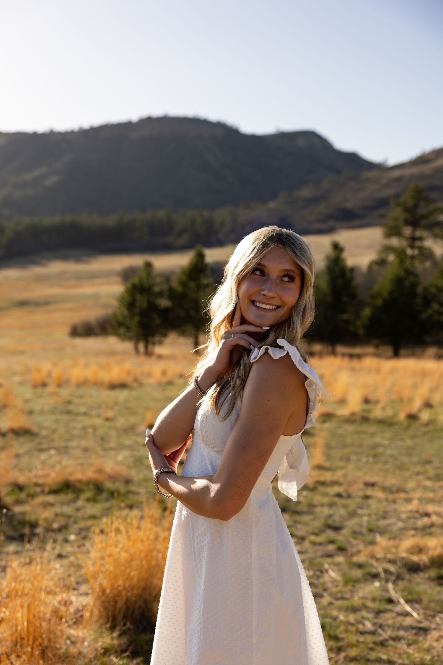 Young woman in white dress smiling over shoulder in golden meadow with mountain backdrop.