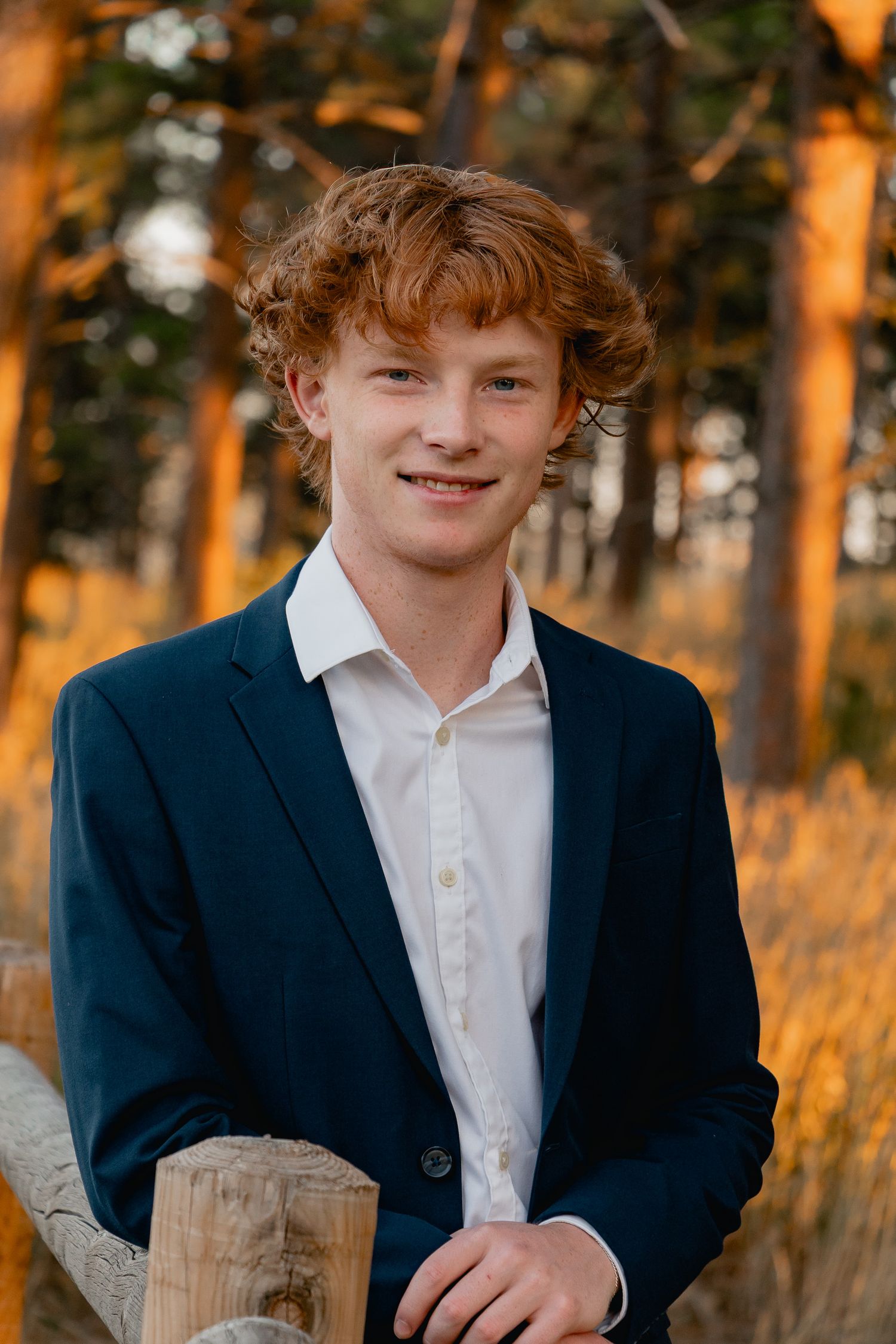 Youth in navy blazer and white shirt shown in stylized portrait against an autumn forest backdrop at golden hour.