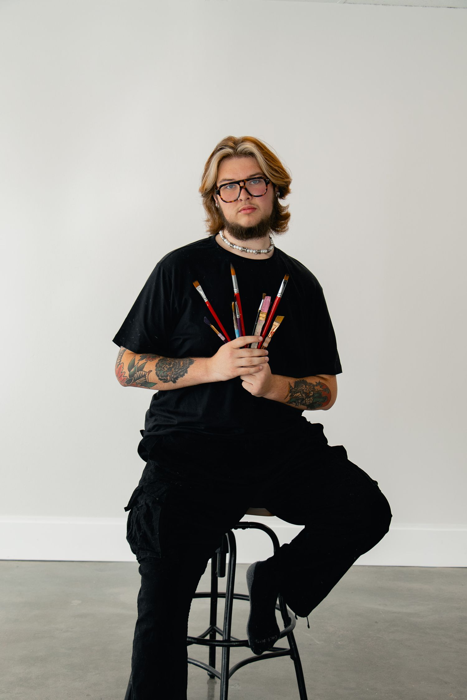 Four progressive shots show someone in black clothing sitting on a stool against a white wall while holding art supplies.