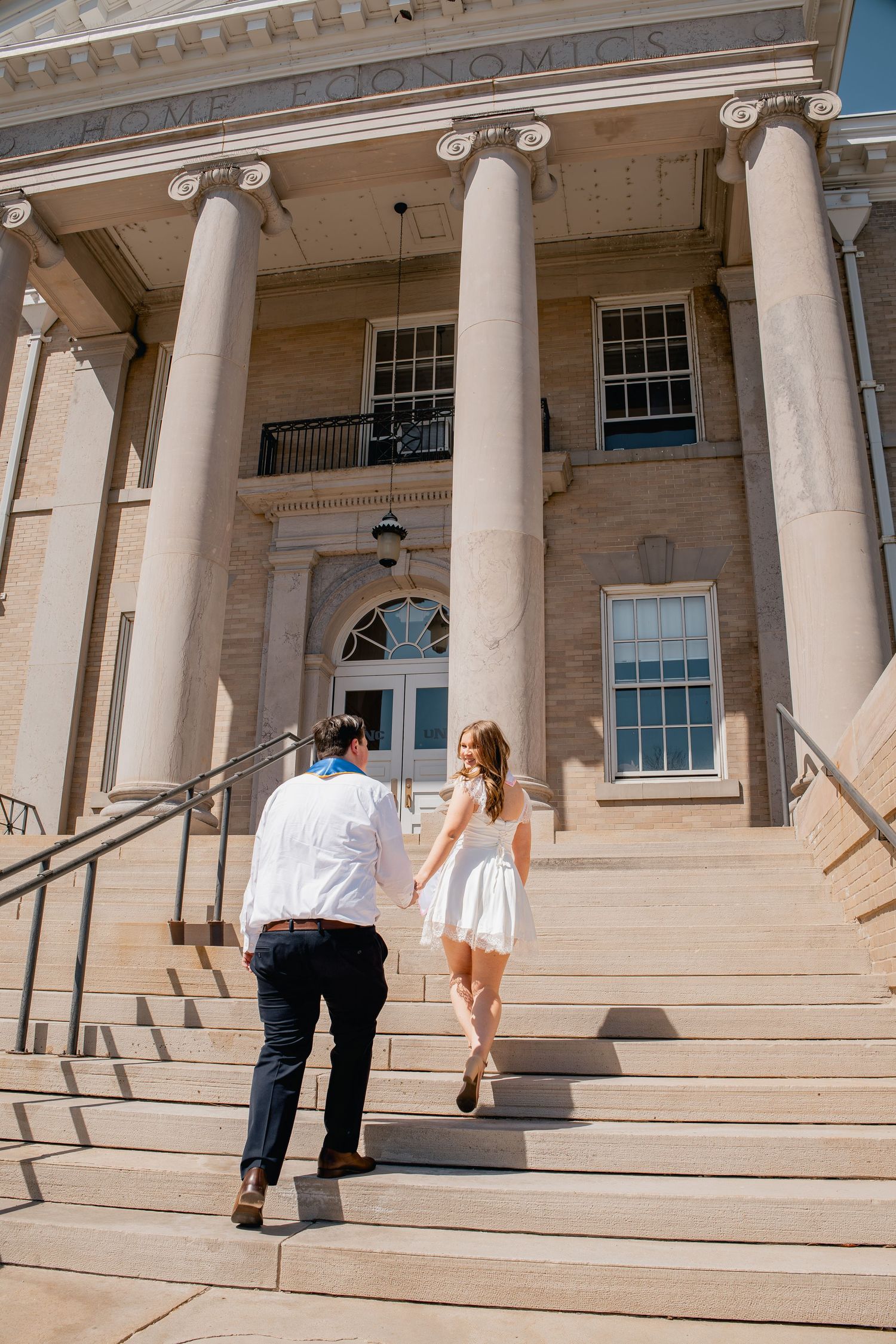 Two people in formal attire walk up the grand stone steps of a neoclassical building with towering Ionic columns.