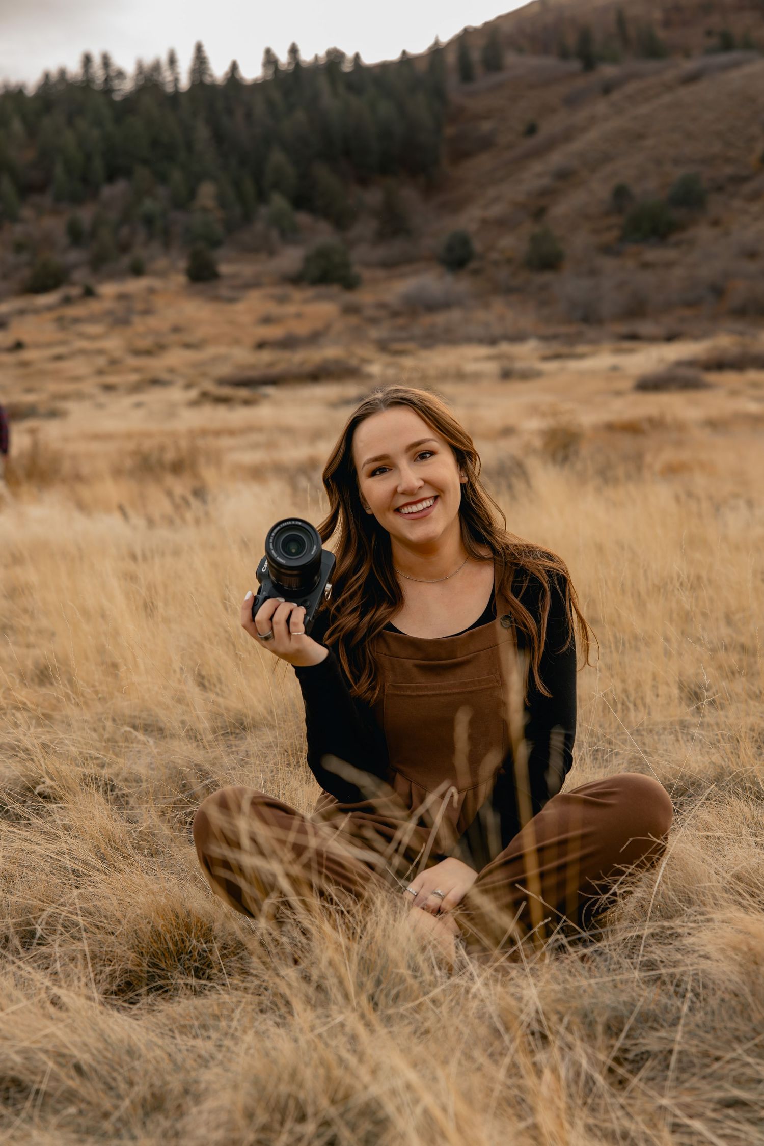 A photographer lying in golden autumn grass holding a camera in a mountain landscape setting.