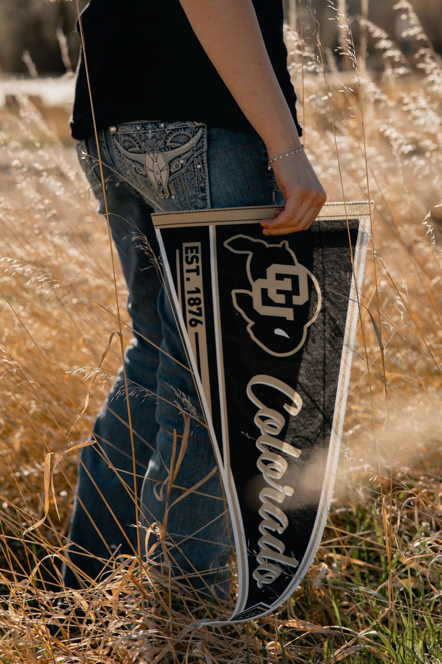 Person holding a University of Colorado Buffaloes pennant EST. 1876 in a golden grass field.