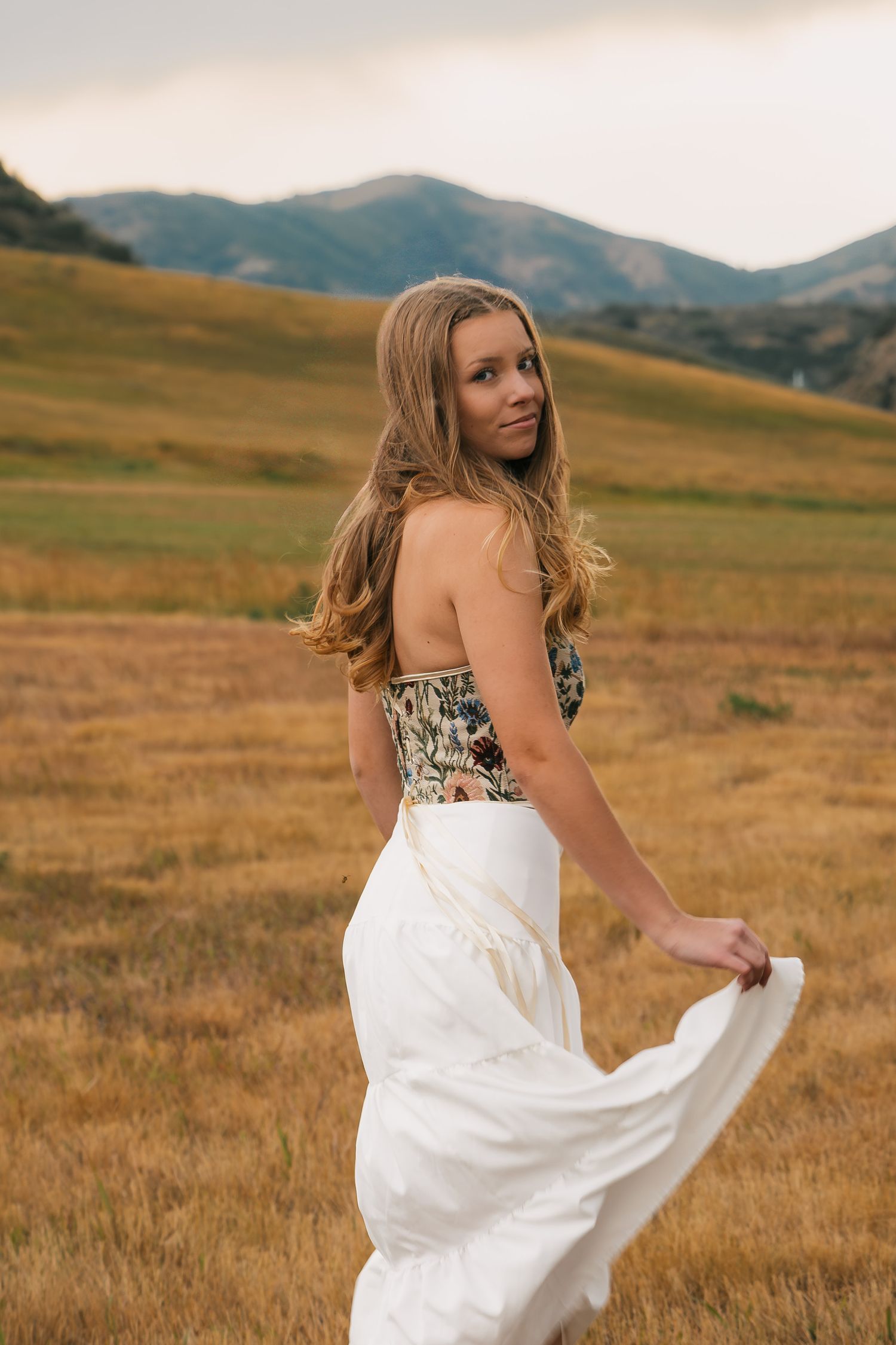 A person in a flowing white skirt and patterned top poses against golden rolling hills and mountains at sunset.