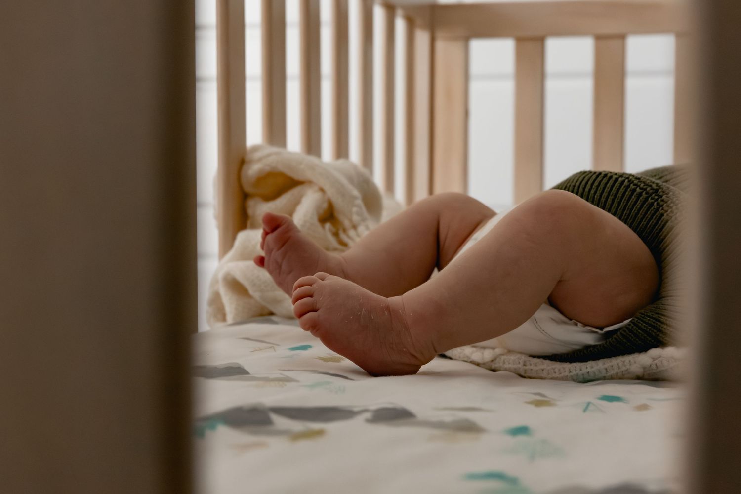 Close-up of a sleeping baby's bare feet and legs in a wooden crib with soft bedding.