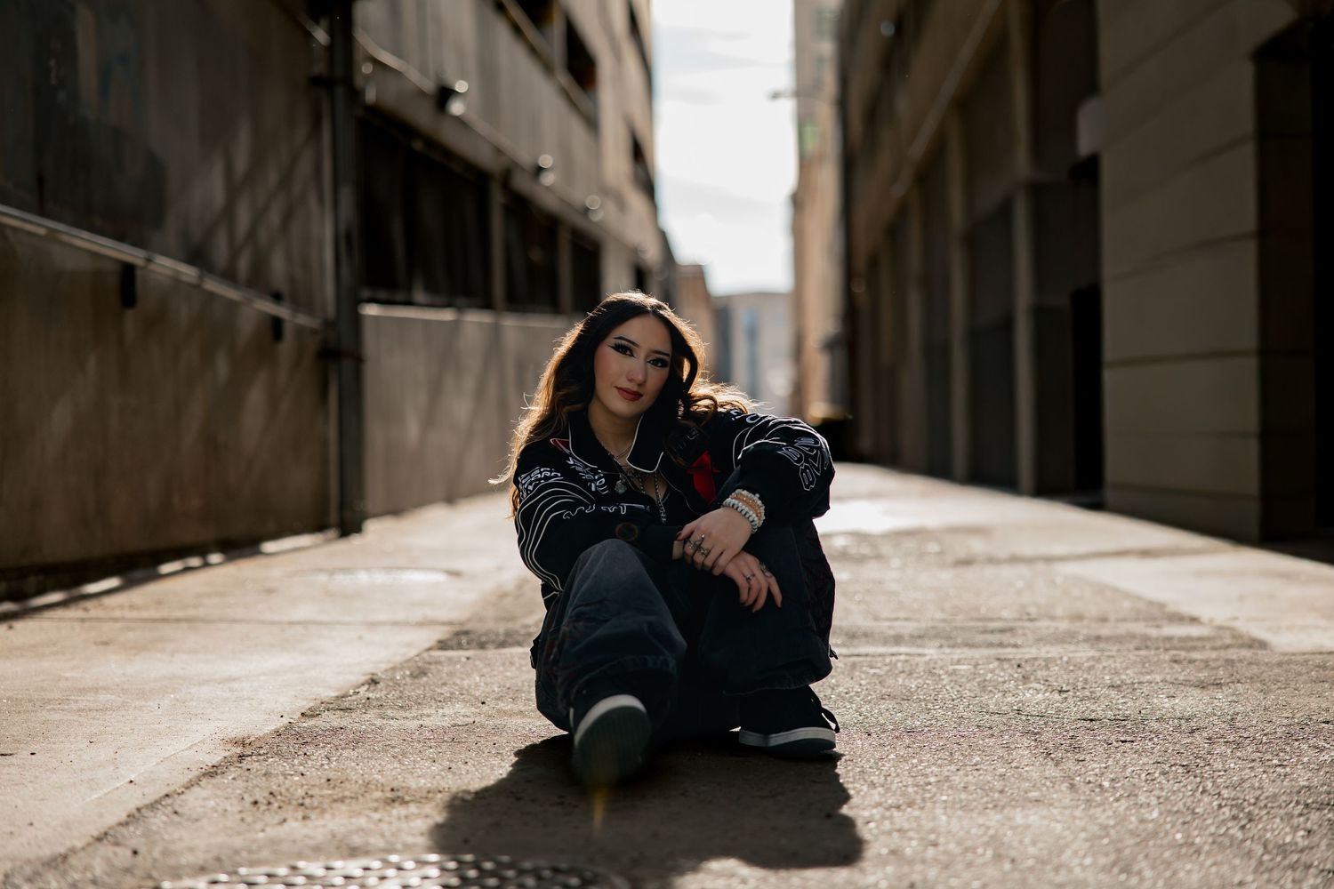 Young woman in dark outfit sitting on urban alley ground between buildings, casual portrait photography.