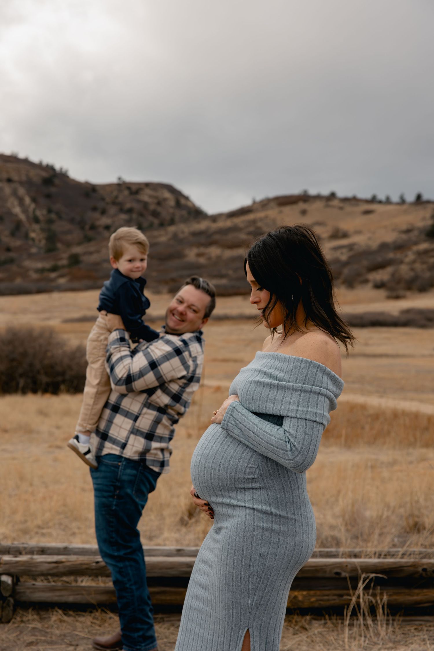 Pregnant woman in gray off-shoulder dress poses outdoors while man holds toddler son in autumn field.