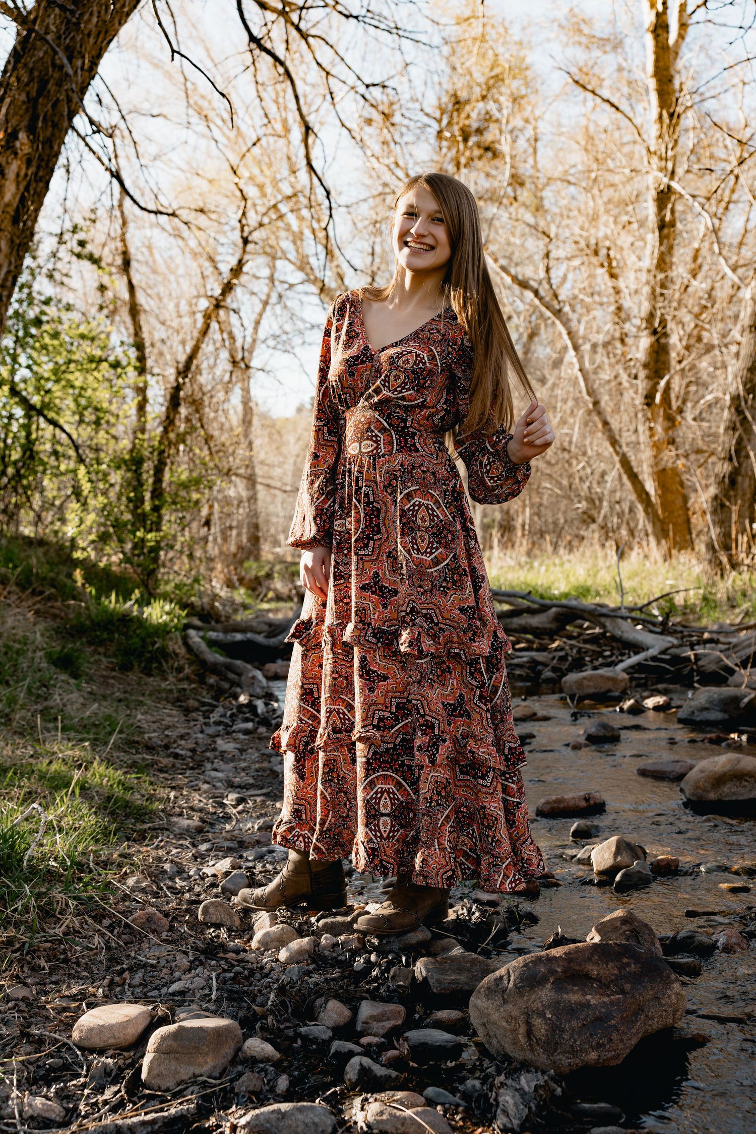 Woman in boho floral maxi dress standing on rocky creek bed surrounded by bare trees in natural outdoor setting.