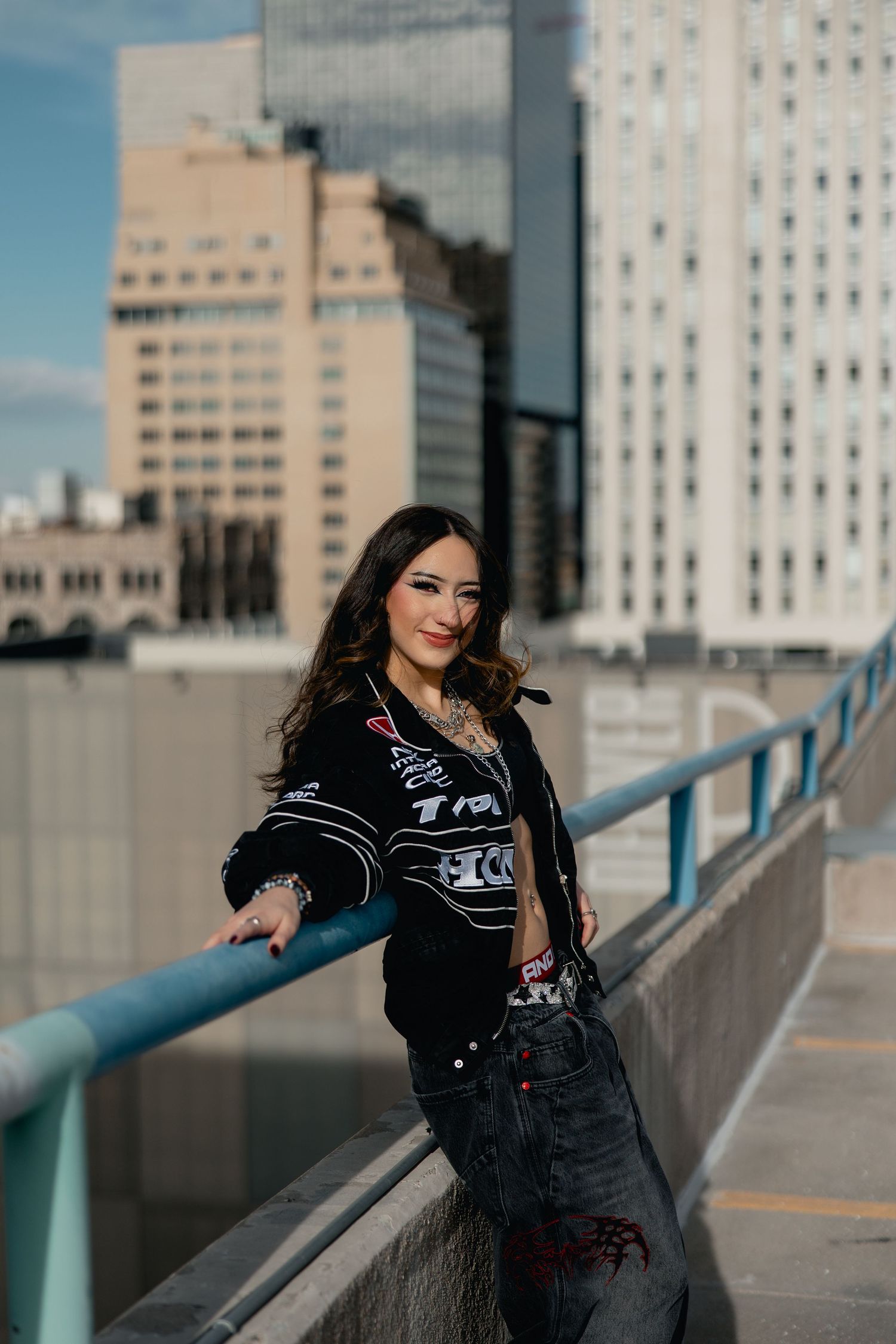 Young woman in black graphic jacket and jeans poses on urban rooftop with city skyline backdrop.