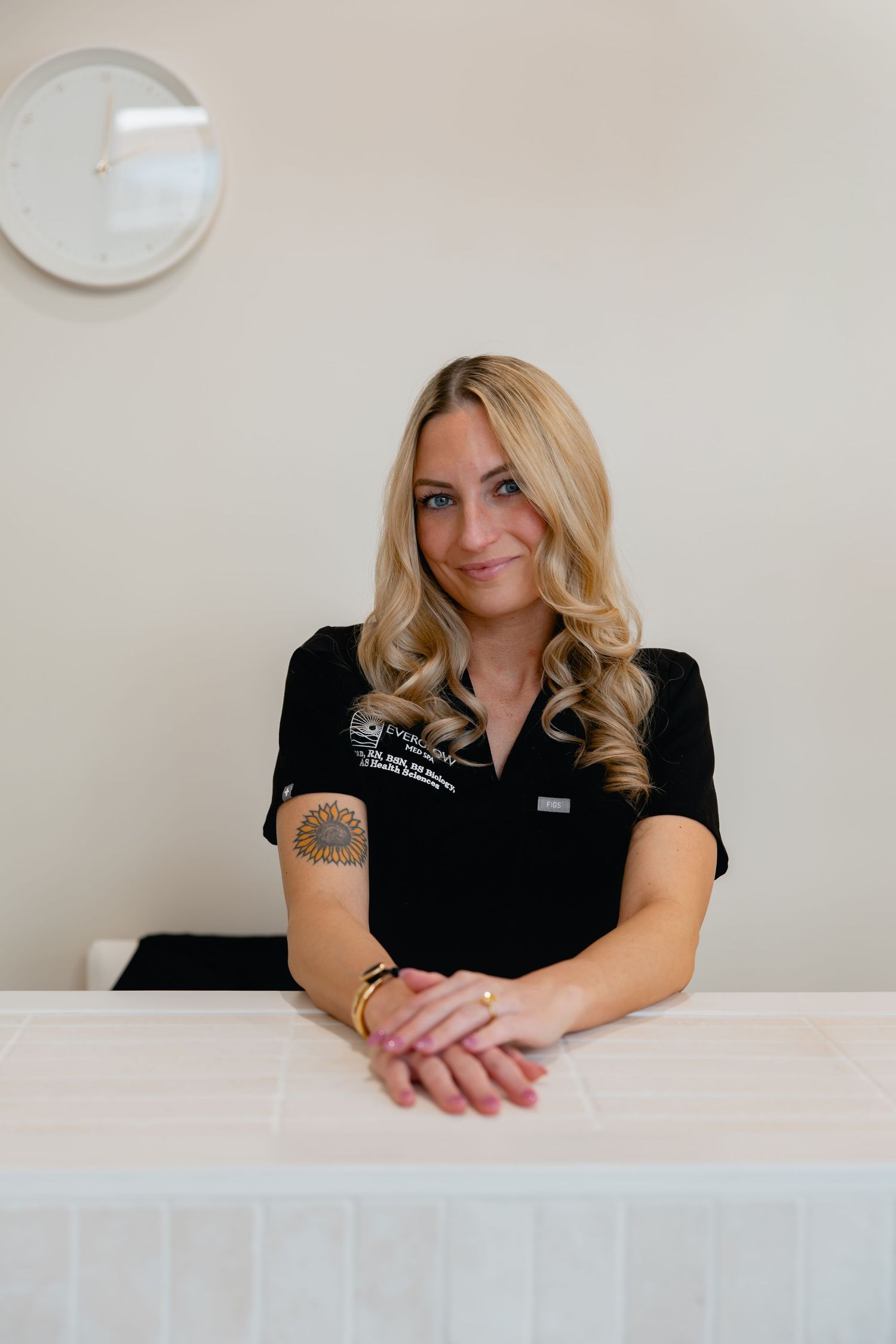 Smiling blonde woman in black uniform with tattoo sits at white reception desk in bright modern spa.