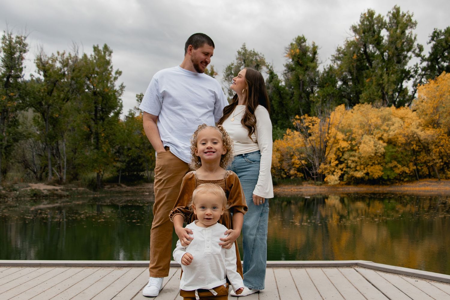 A family enjoys a playful moment together on a wooden dock by an autumn lake with golden trees in the background.