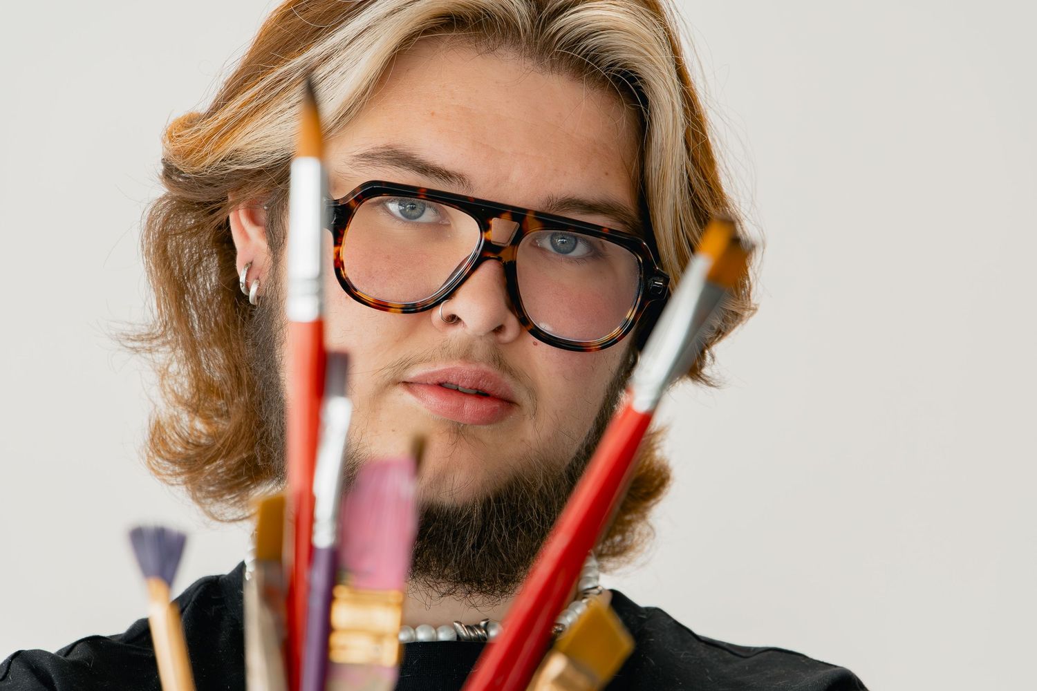 Creative professional wearing thick-rimmed glasses holds art supplies and brushes against white background.