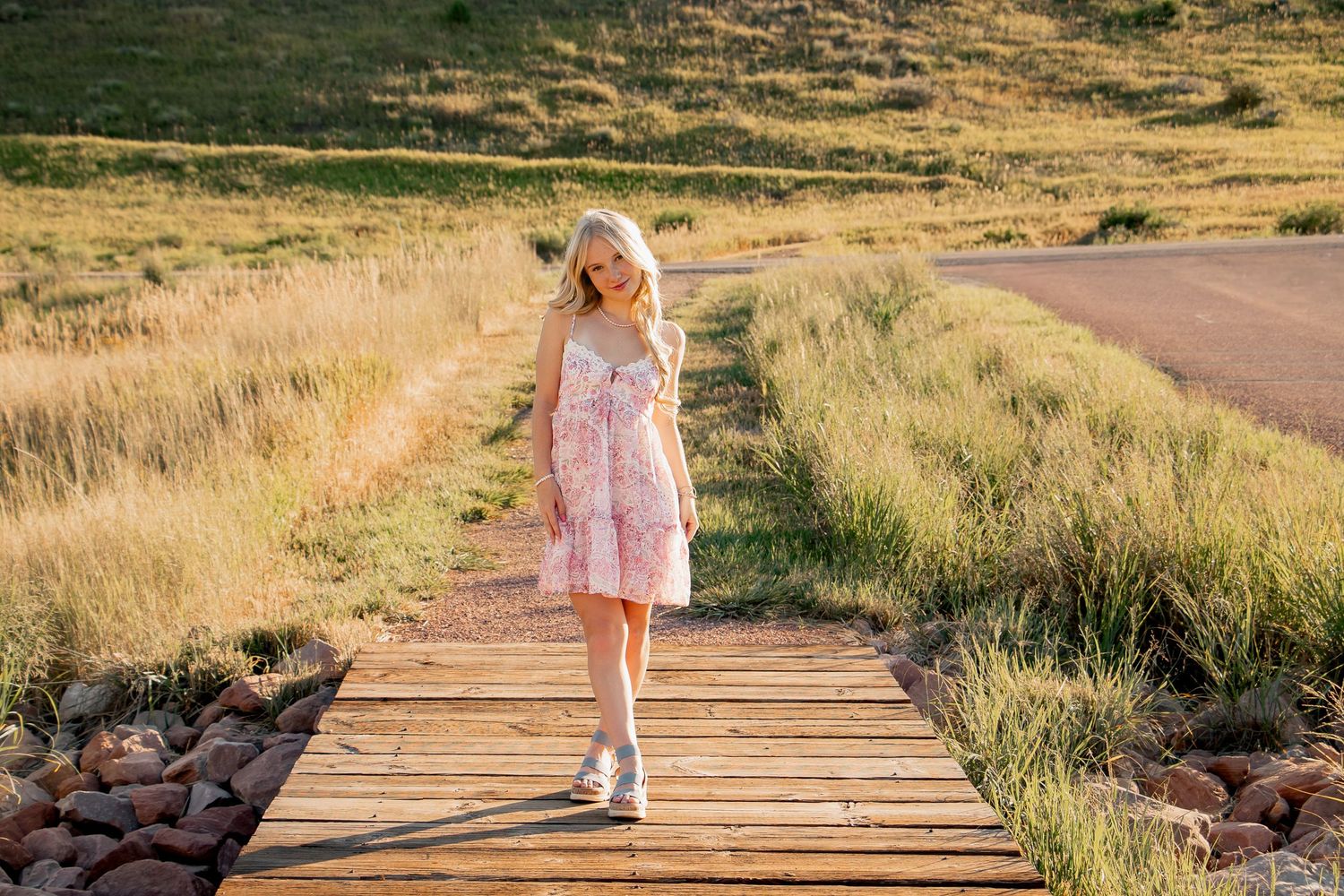 A figure stands on a wooden walkway surrounded by golden grasses during sunset on a summer evening.