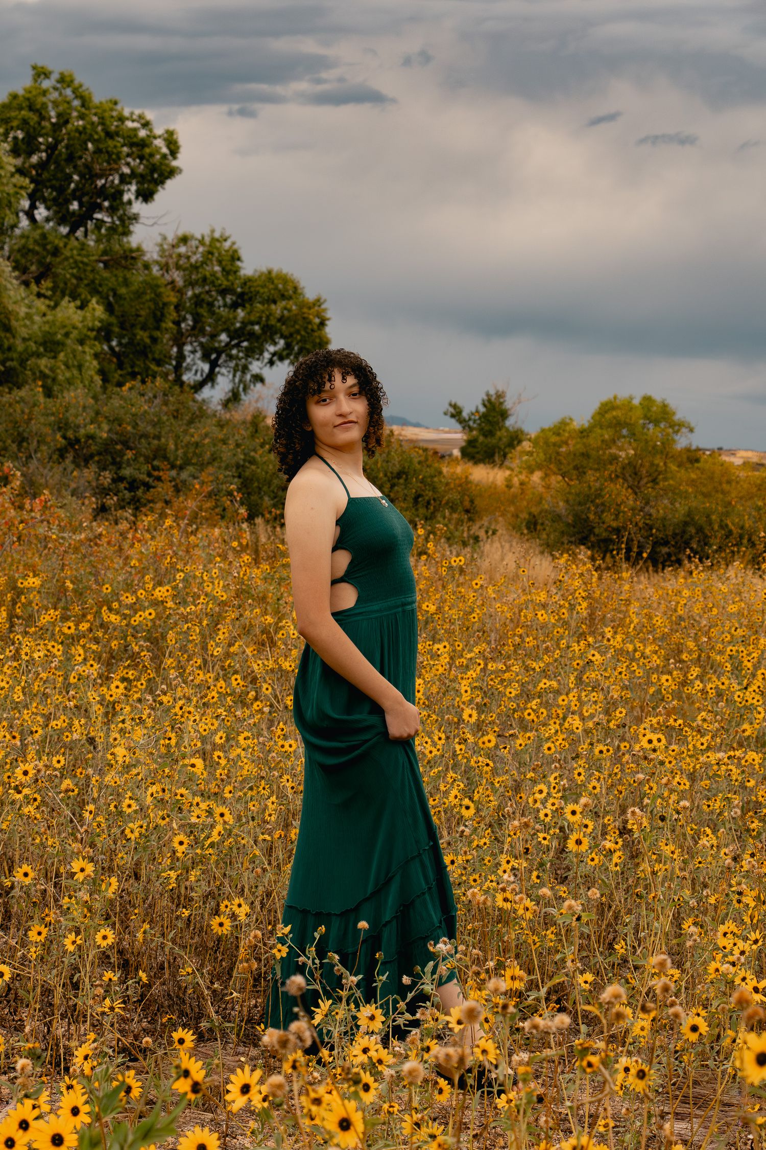 A figure in an emerald green dress stands among vibrant yellow wildflowers in a summer meadow under cloudy skies.