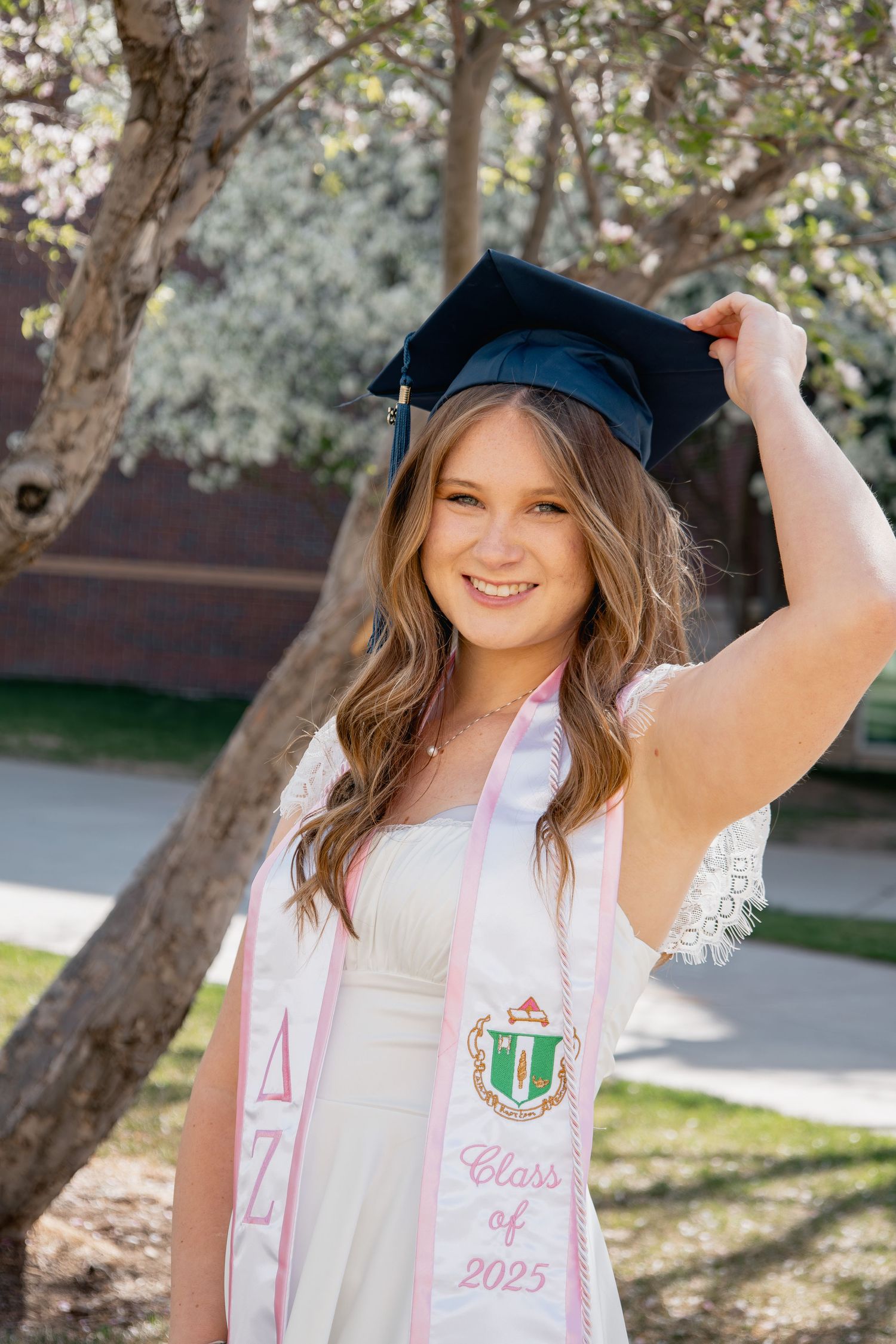 A graduate in a cap and gown poses outdoors with a class of 2023 graduation sash and sorority stole during springtime.
