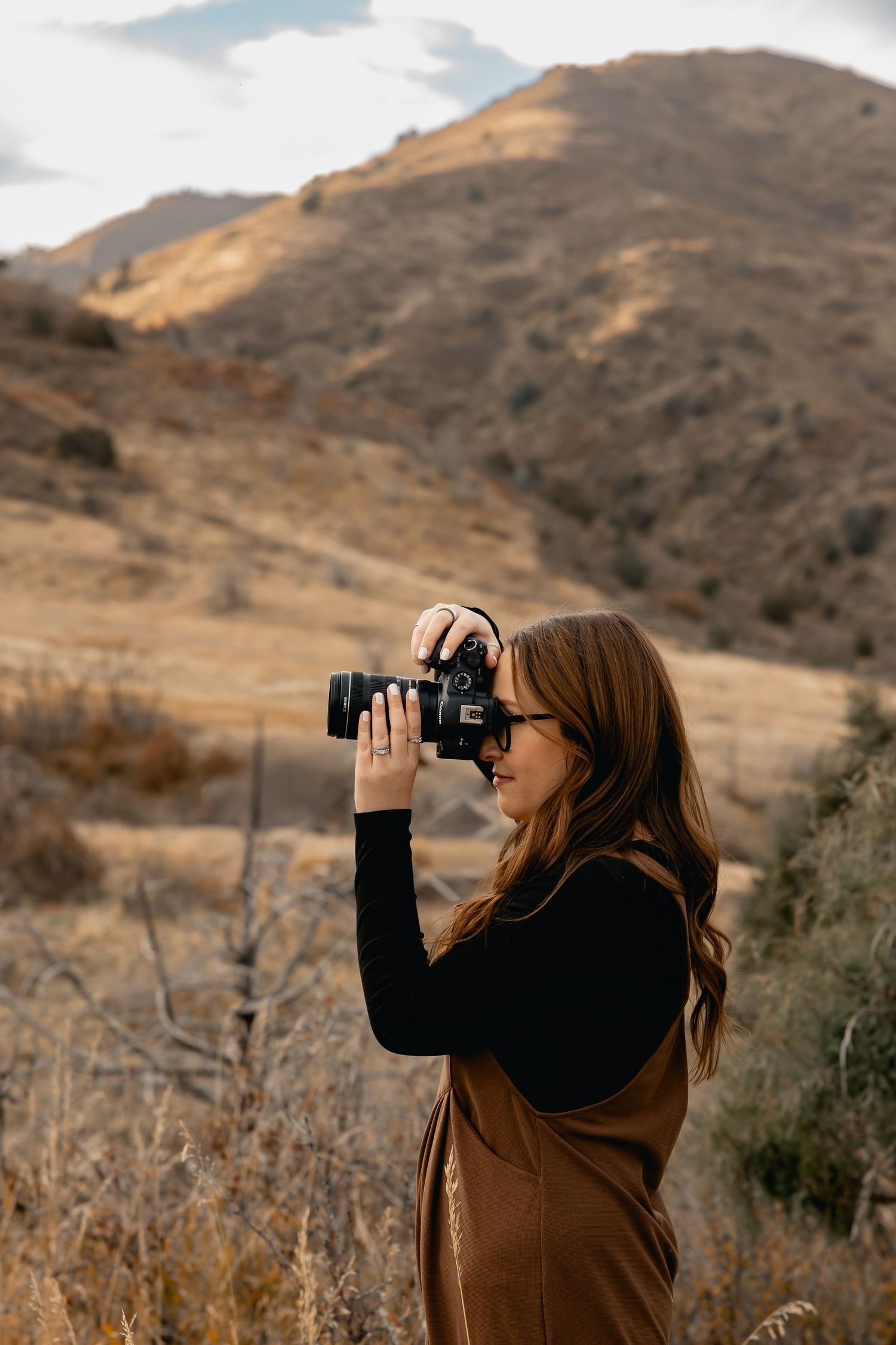 Woman photographer capturing mountain landscape with DSLR camera, wearing brown vest outdoors in autumn hills.