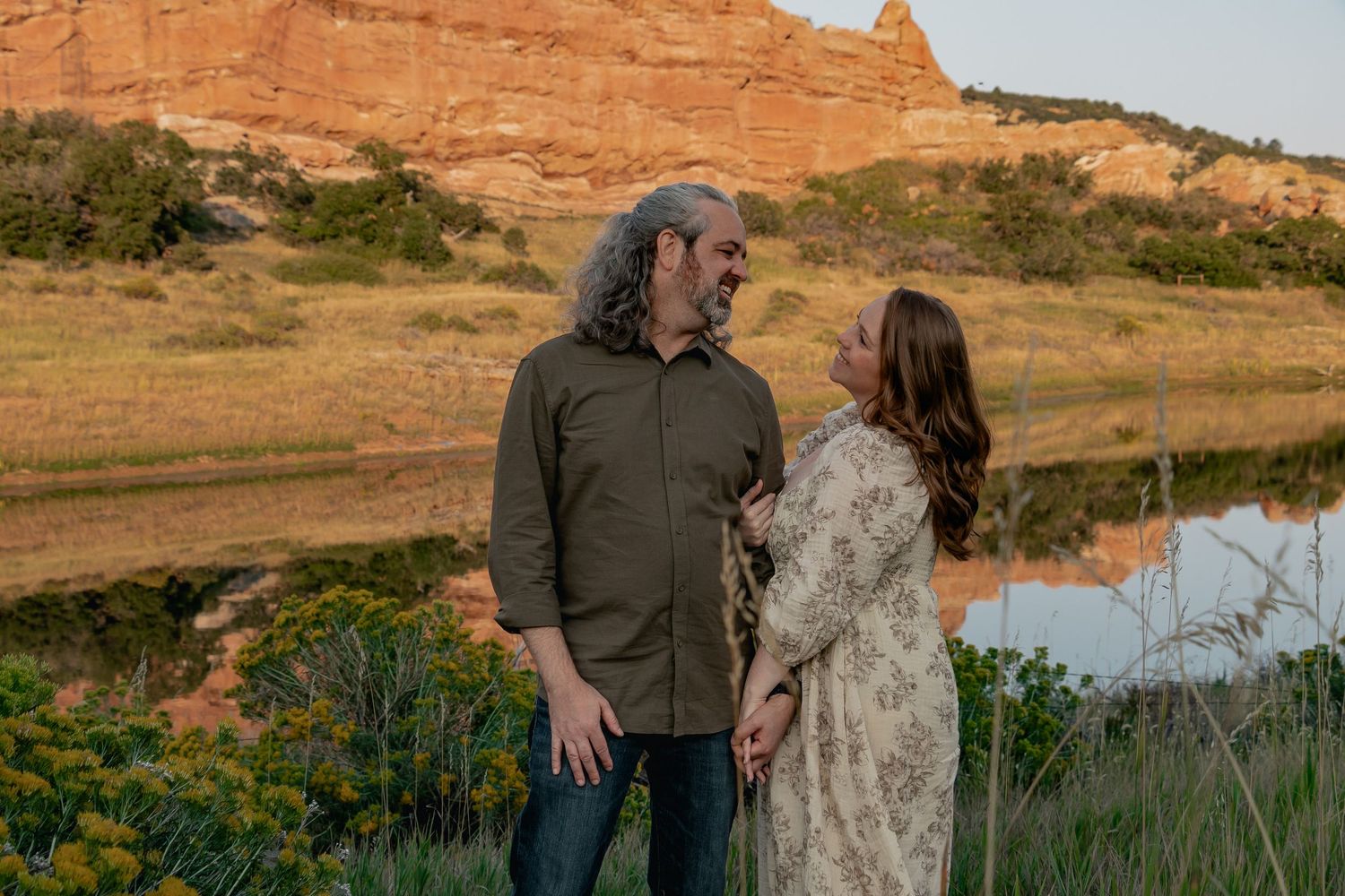 Two people share a tender moment against a backdrop of red rock formations and water at sunset in a desert landscape.