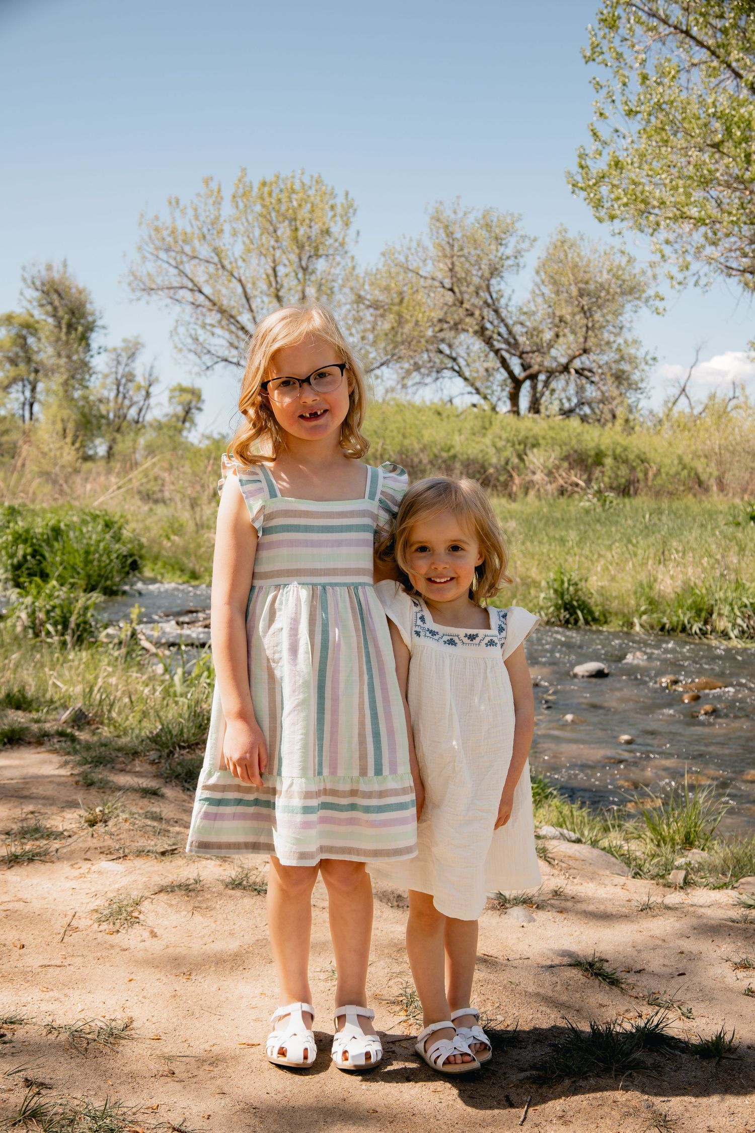 Two children wearing summer dresses stand together on a dirt path with trees and natural scenery in the background.