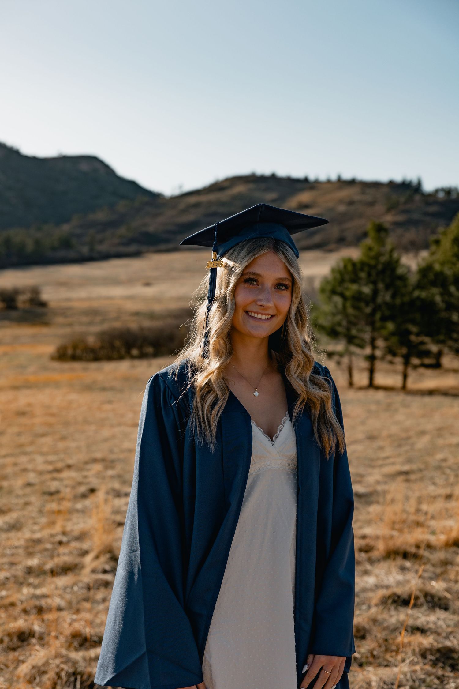 Smiling graduate in blue cap and gown posing outdoors in a golden field with hills in the background.