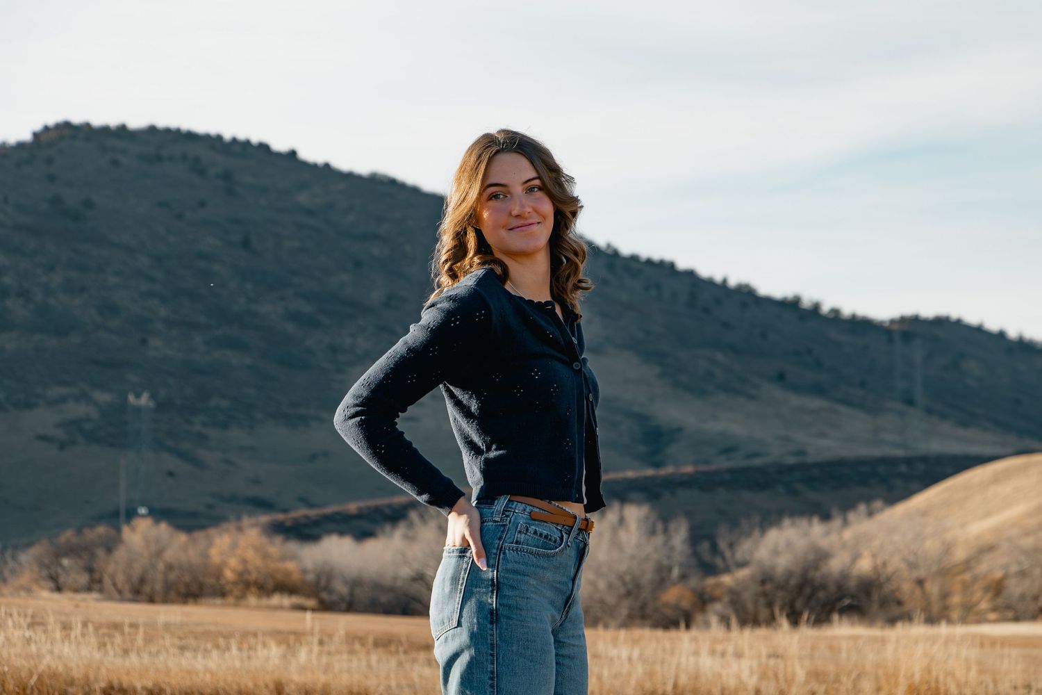 A person in a black sweater and jeans stands in a golden field with mountains in the background.