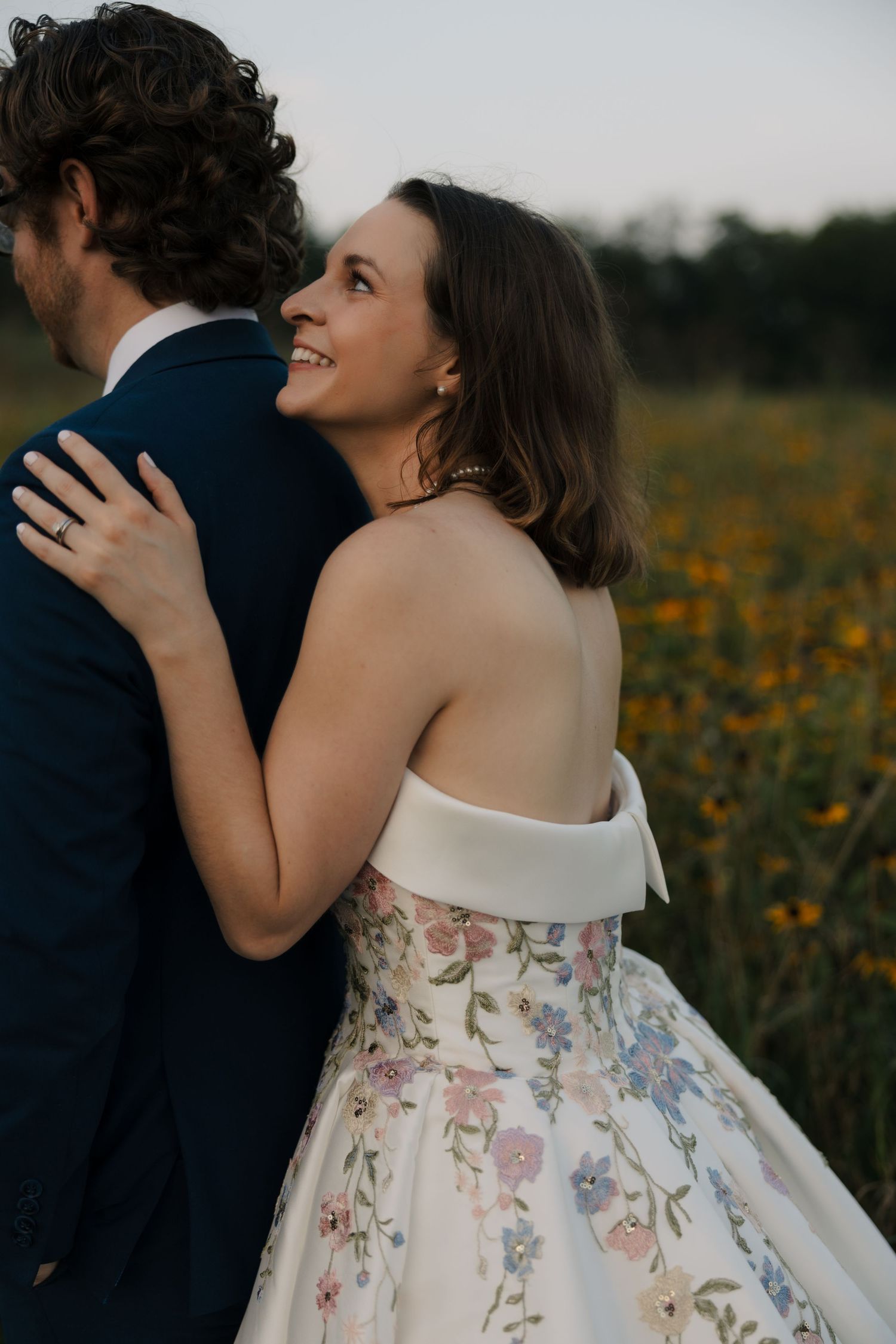 Smiling bride in floral off-shoulder gown embraces groom in navy suit in sunflower field at sunset.