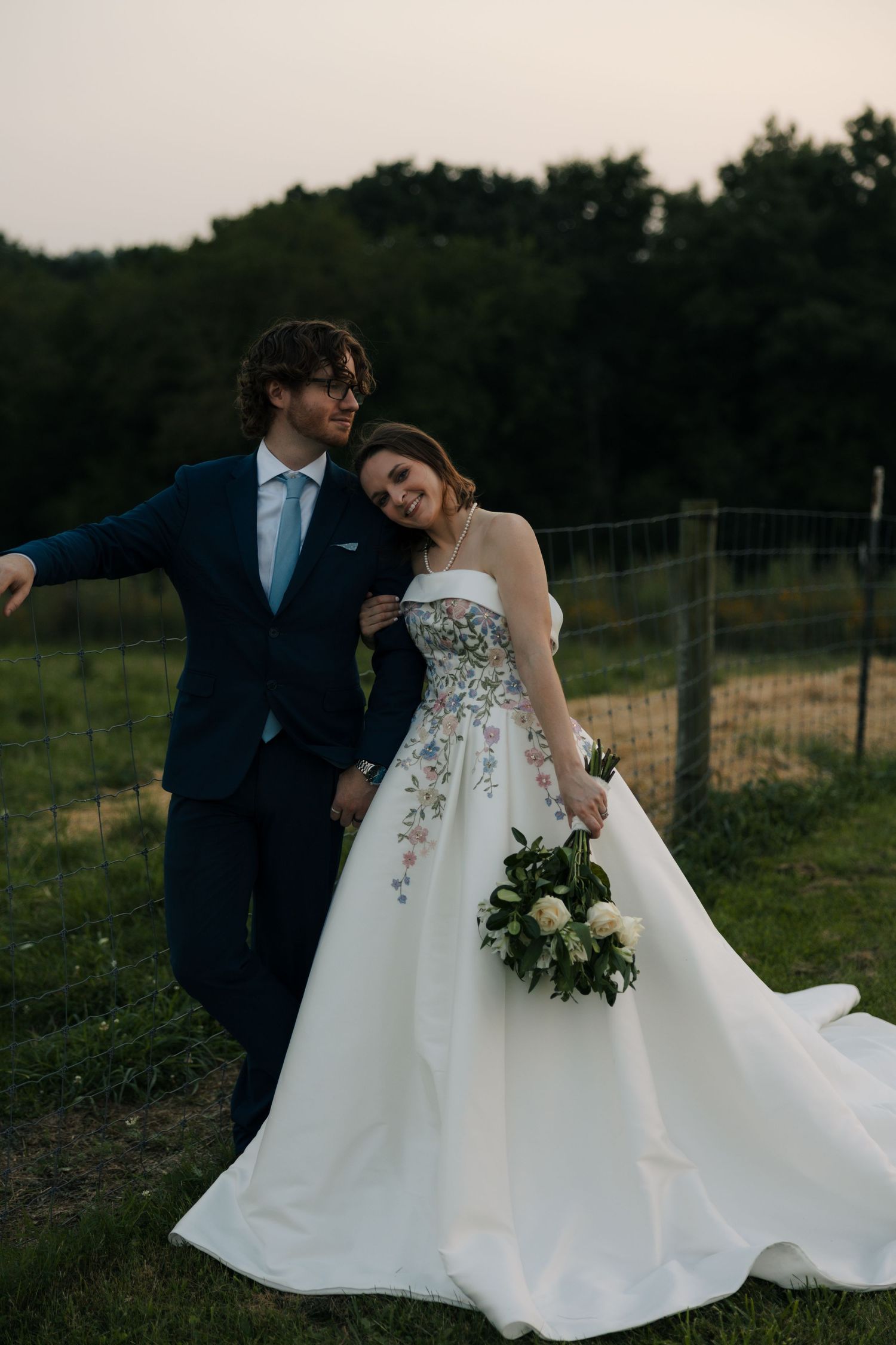 Groom in navy suit embraces bride in floral strapless gown near rustic fence at golden hour.