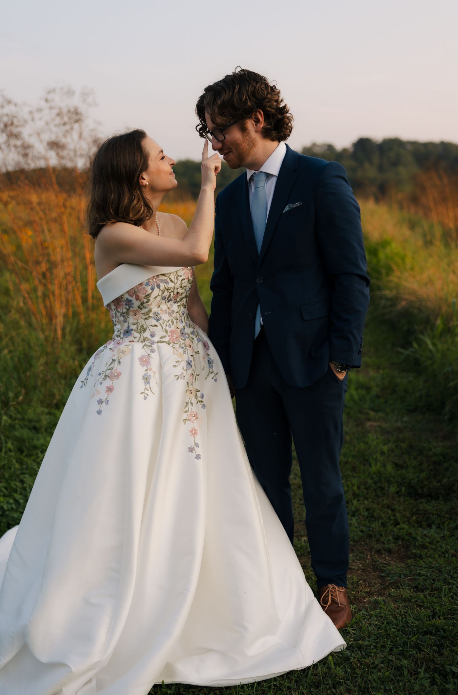 Bride in floral ball gown touches grooms nose as they smile at each other in golden hour field.