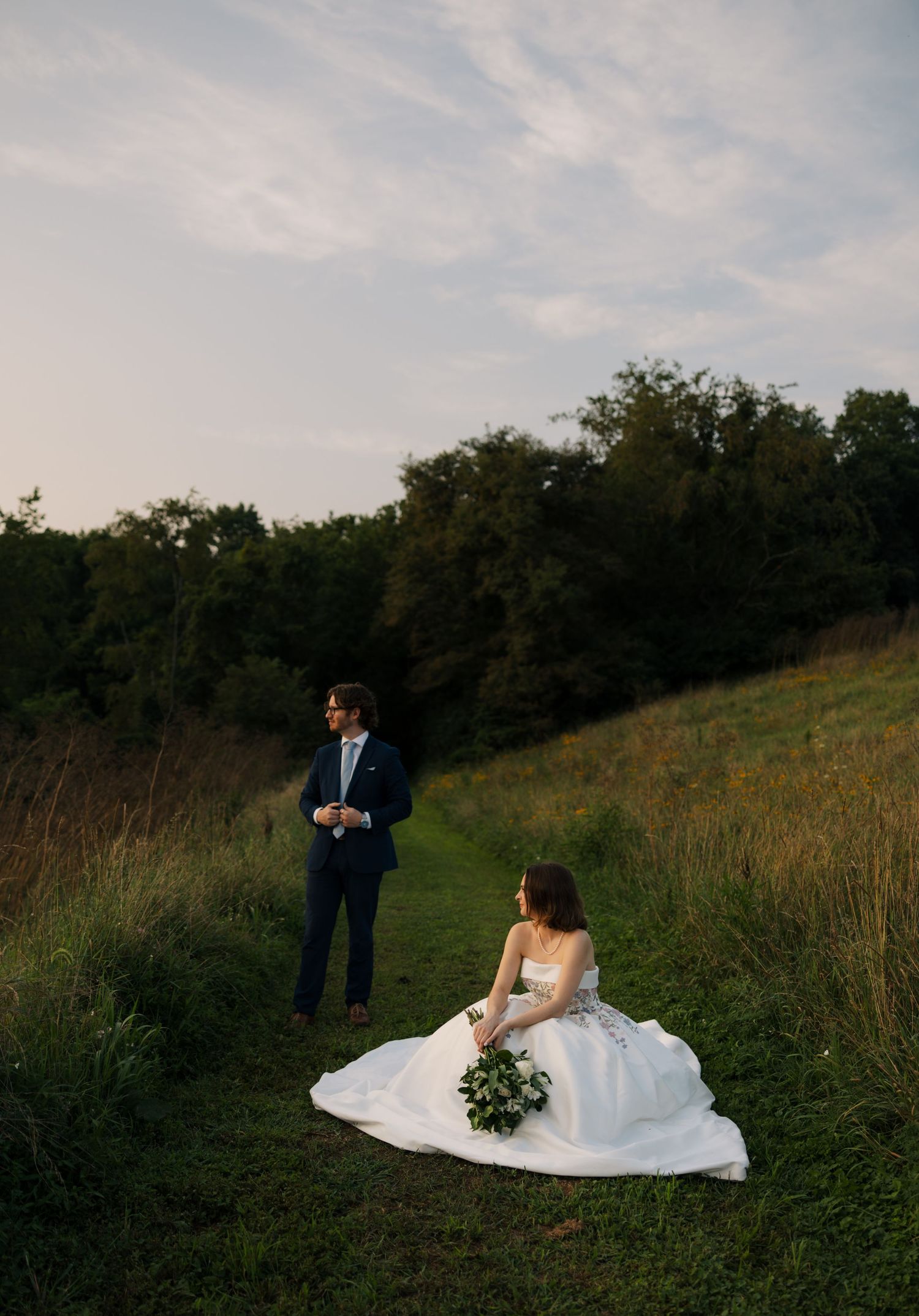 Bride in white floral gown sits on green hillside holding bouquet while groom in navy suit stands nearby at dusk.