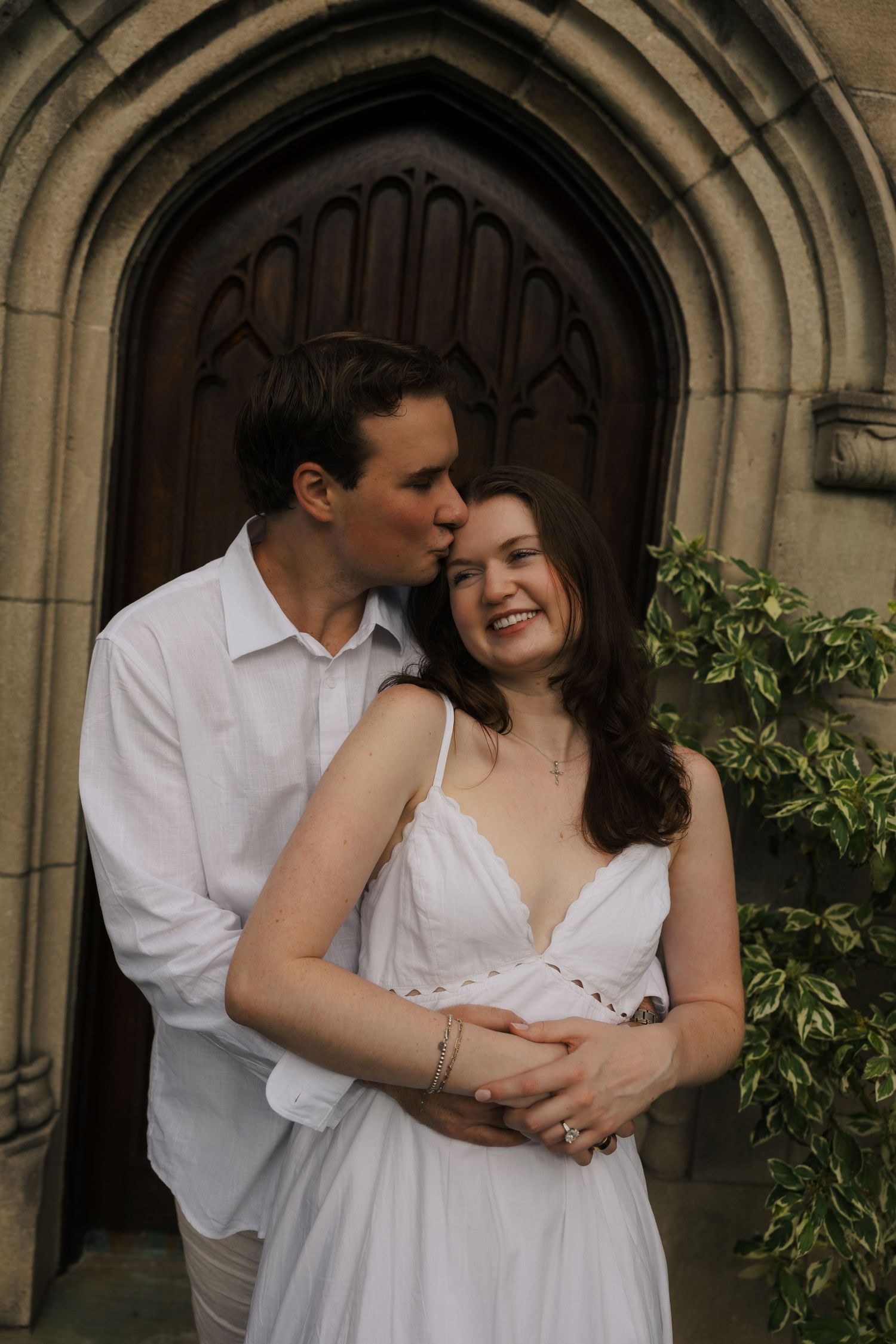 Smiling couple embracing before ornate Gothic wooden church door with ivy backdrop.