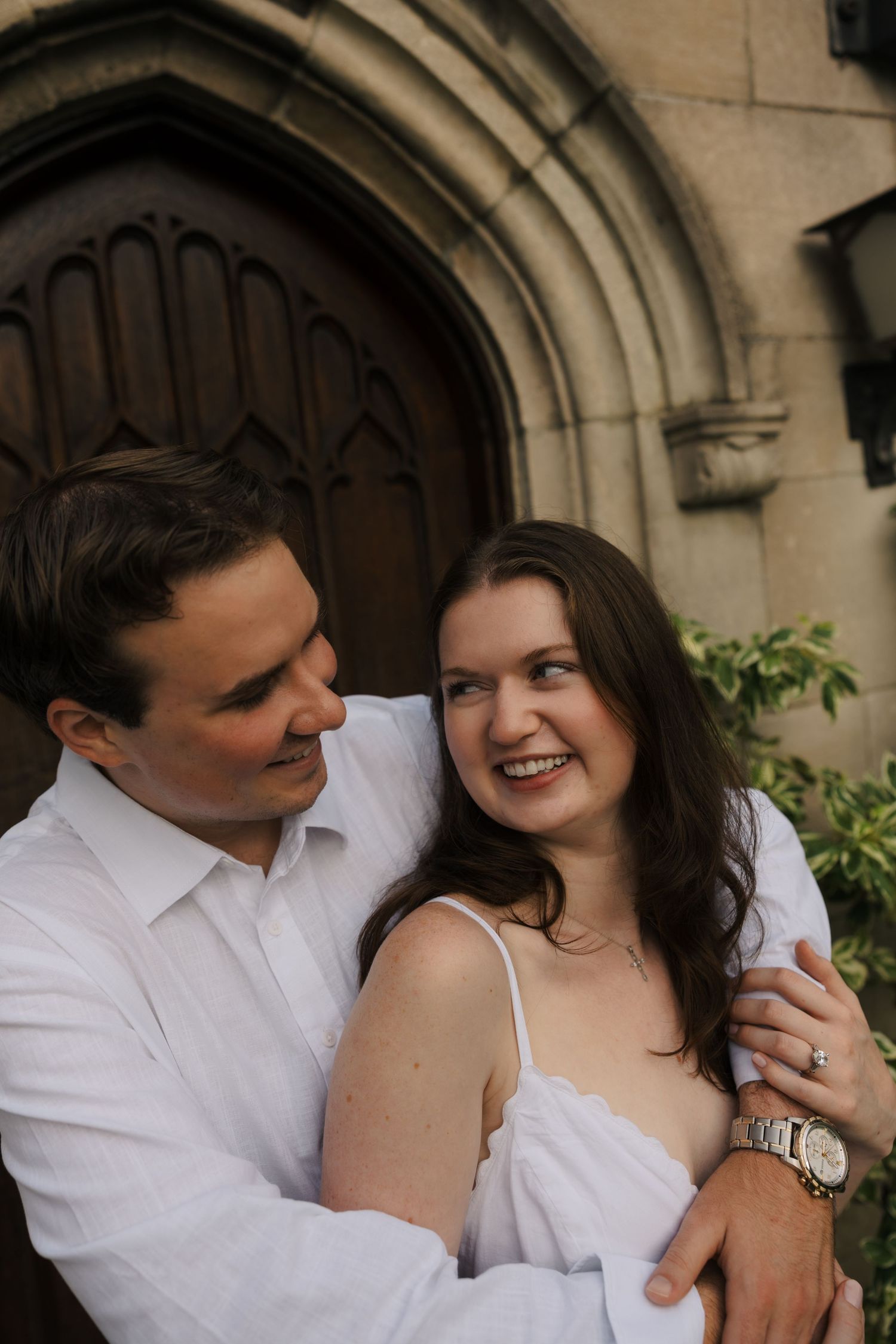 Happy engaged couple smiling together in front of ornate wooden church doors, woman wearing ring.