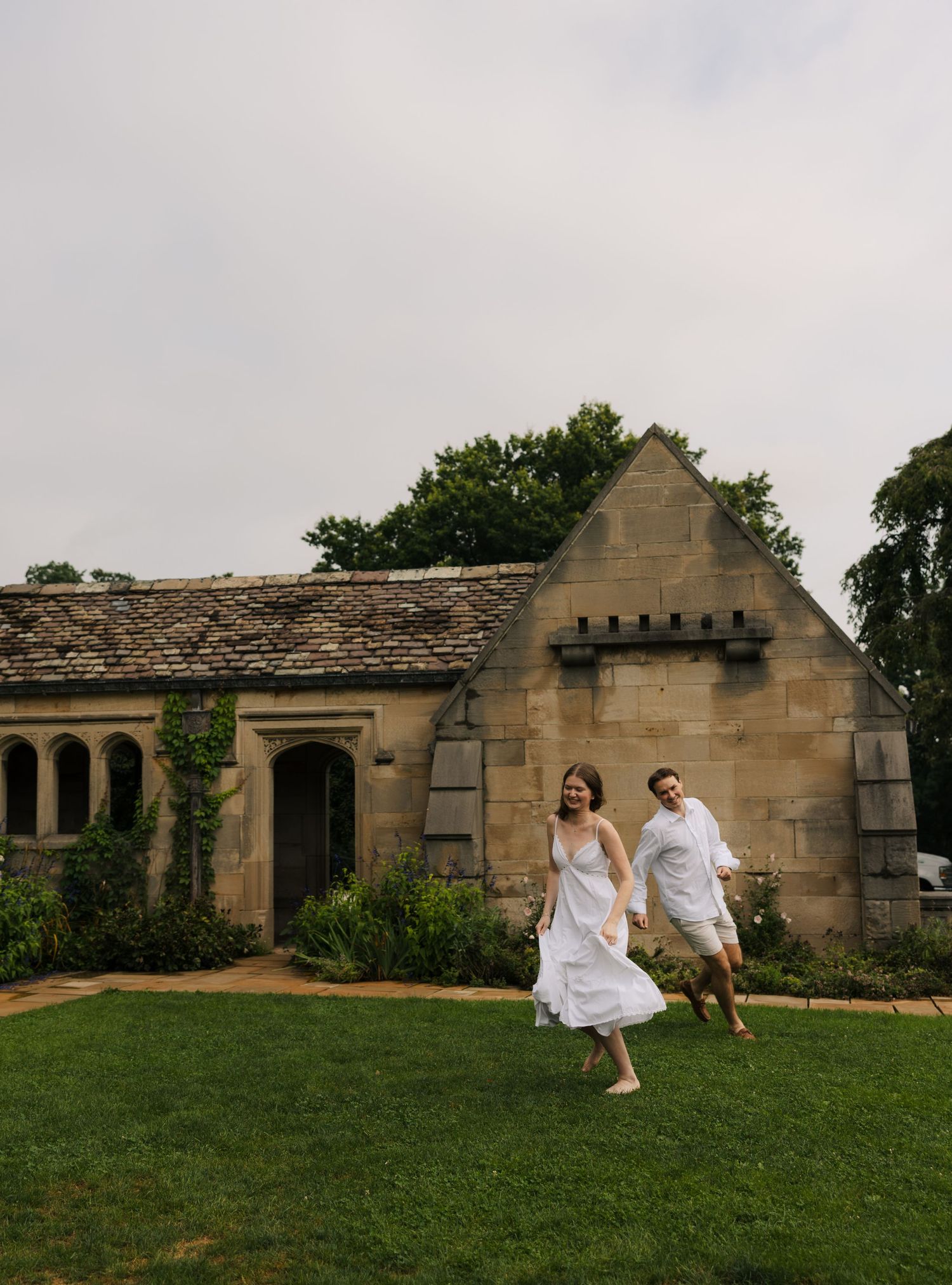 Happy couple in white outfits runs hand-in-hand across lawn before charming stone chapel.