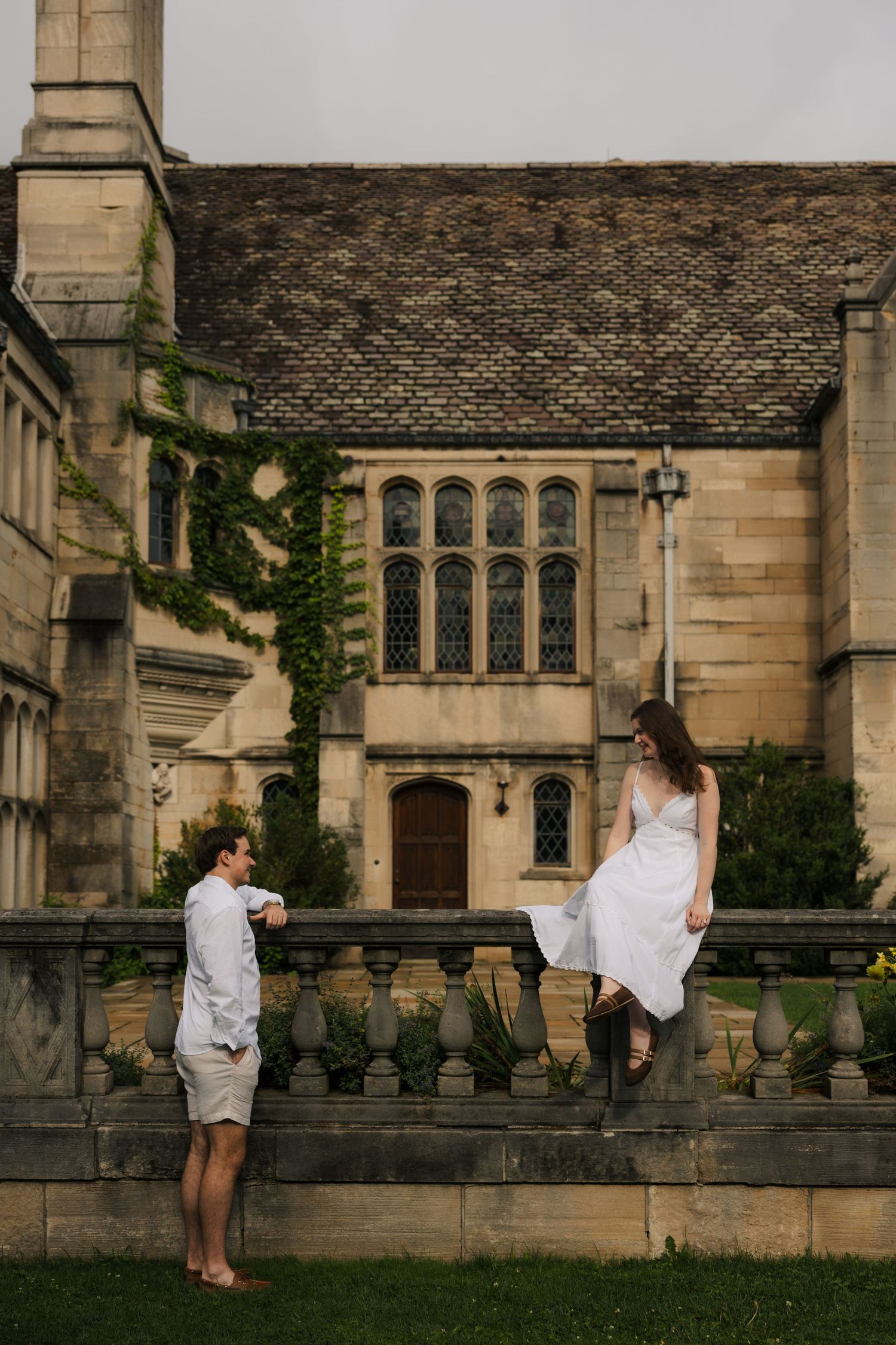A woman in a white dress sits atop a stone balustrade while a man looks up at her by a grand manor.